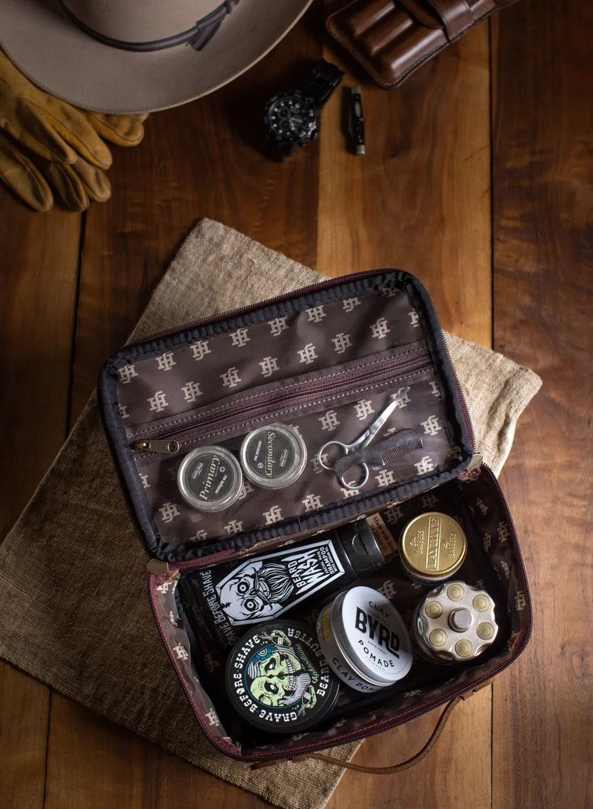 Open cosmetic bag containing grooming products such as beard wax, clay pomade, and other grooming items on a wooden table with a towel underneath.