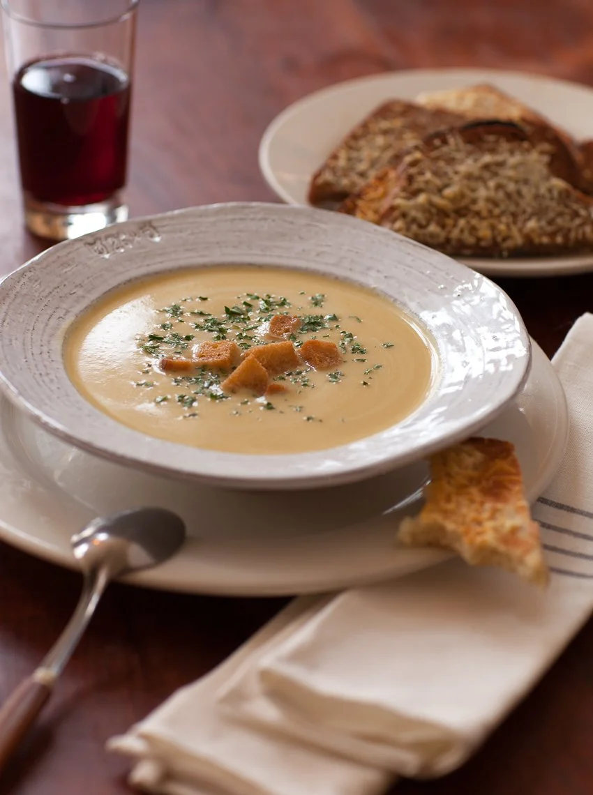 Bowl of creamy soup garnished with herbs and croutons, served with a slice of pizza, a glass of red beverage, and a plate of garlic bread on a wooden table.