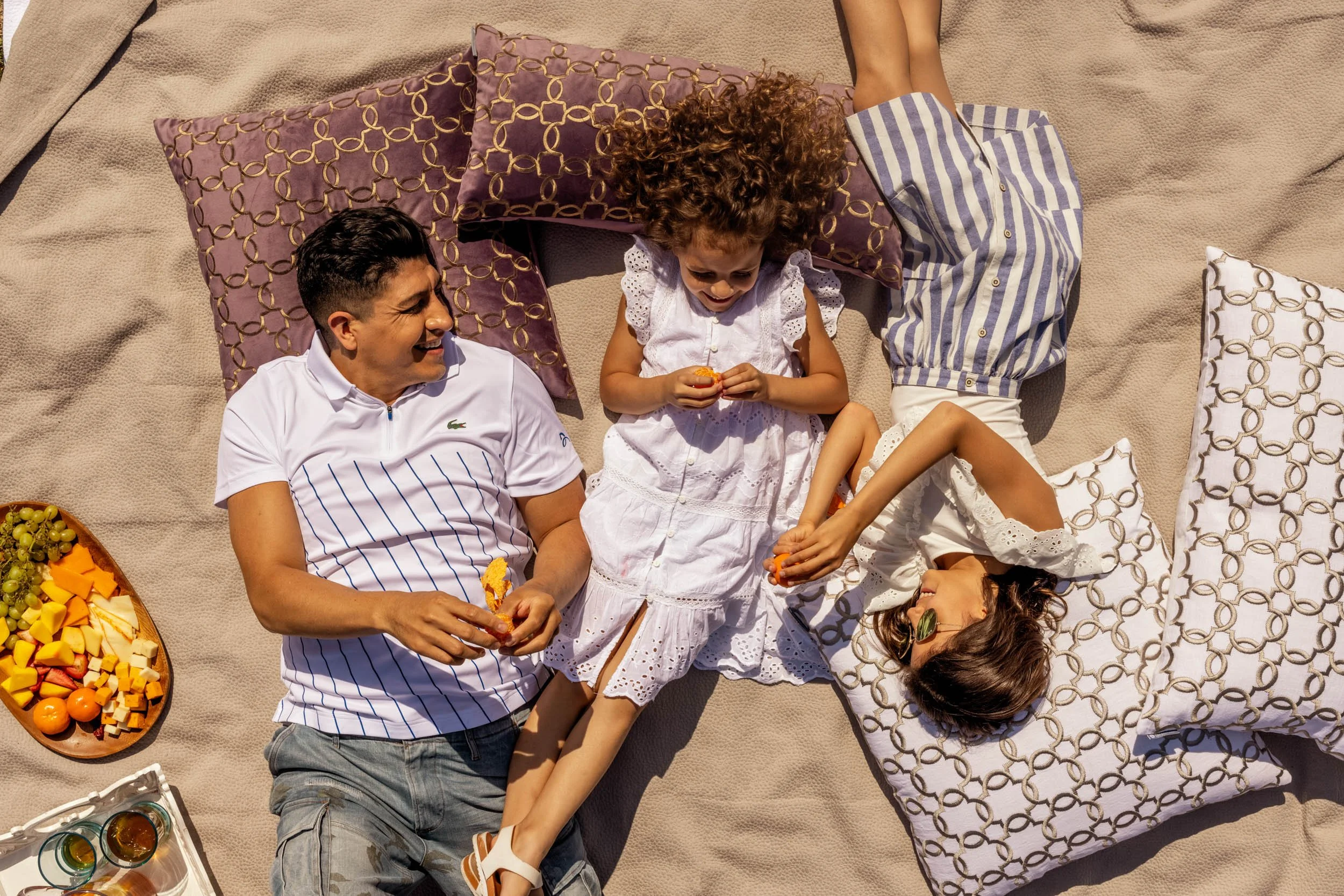 A family of four lying on a beige blanket outdoors, with a girl in a white dress and two boys, one in a striped shirt, surrounded by pillows and food, enjoying a picnic together.