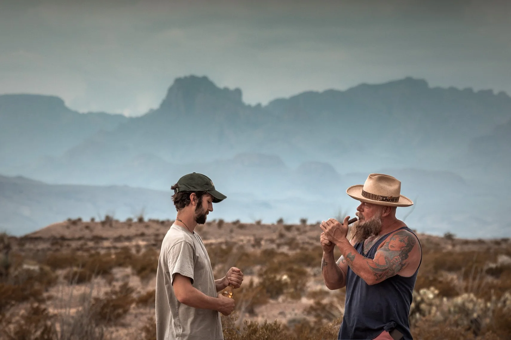 Two men standing in a desert landscape with mountains in the background. One man, younger, with a beard, wearing a gray cap and t-shirt, holding a bottle. The other man, older, with tattoos, a gray beard, wearing a wide-brimmed hat and sleeveless shi