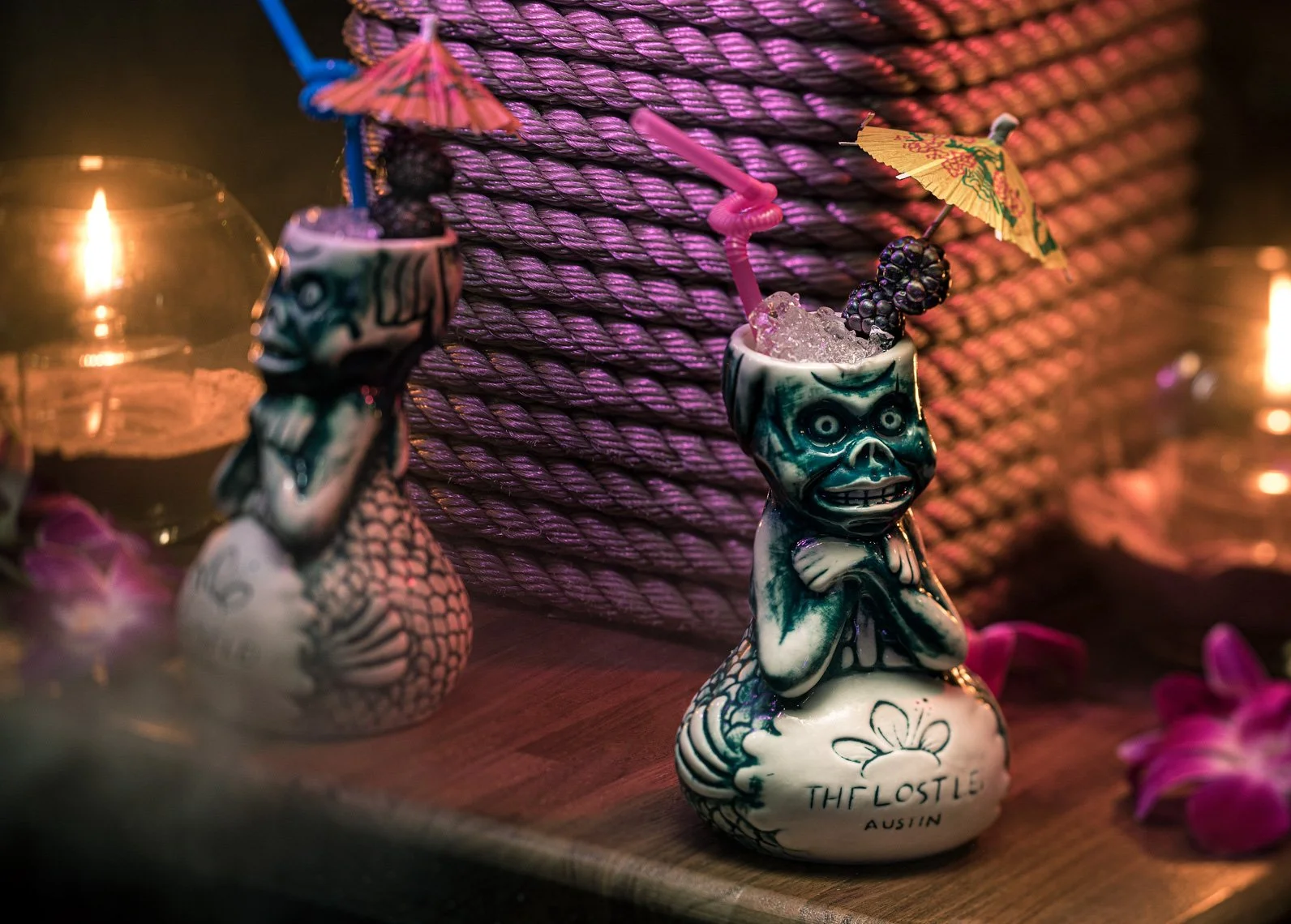 Colorful tiki mugs shaped like skulls with drinks inside, decorated with miniature umbrellas and fruit, placed on a wooden surface with a large woven basket in the background, lit with warm ambient light.