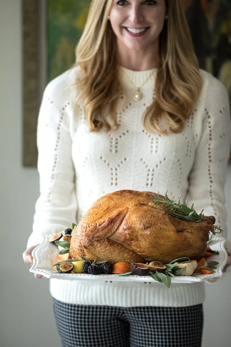 A woman in a white sweater holds a large roasted turkey on a platter decorated with grapes and herbs.
