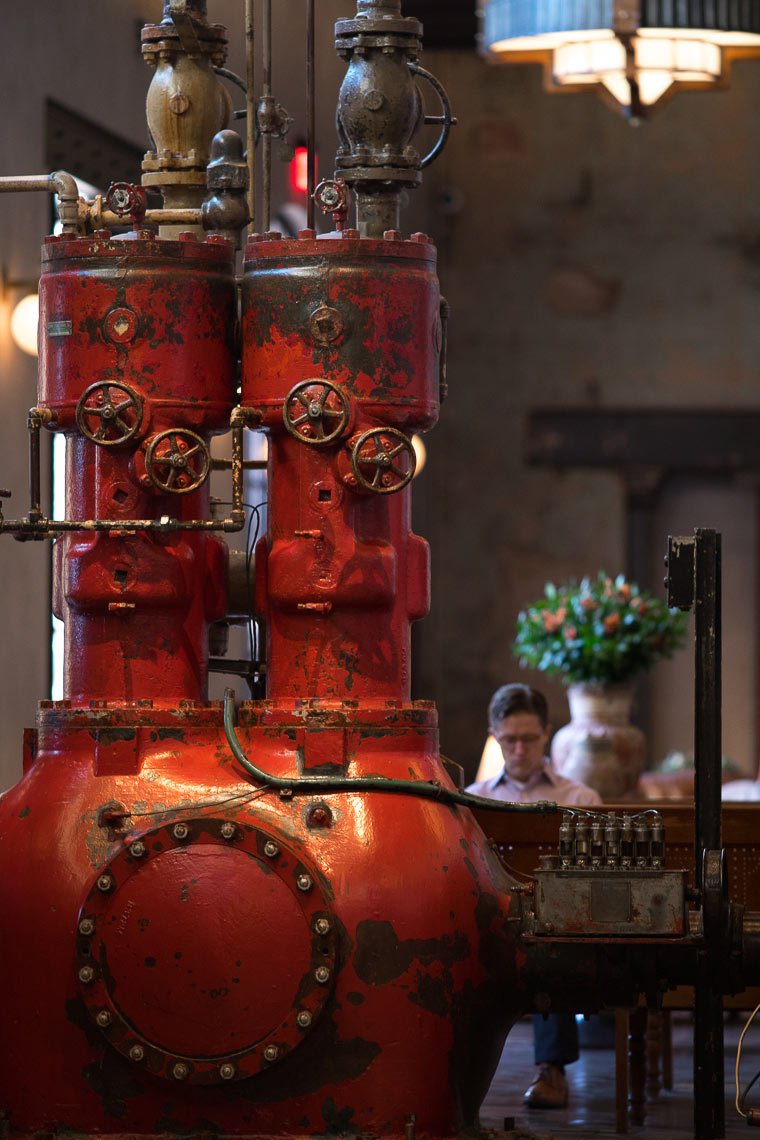 Old red industrial machine, possibly a boiler or engine, with pipes, valves, and gauges, in a vintage-style interior with a person sitting at a table in the background.