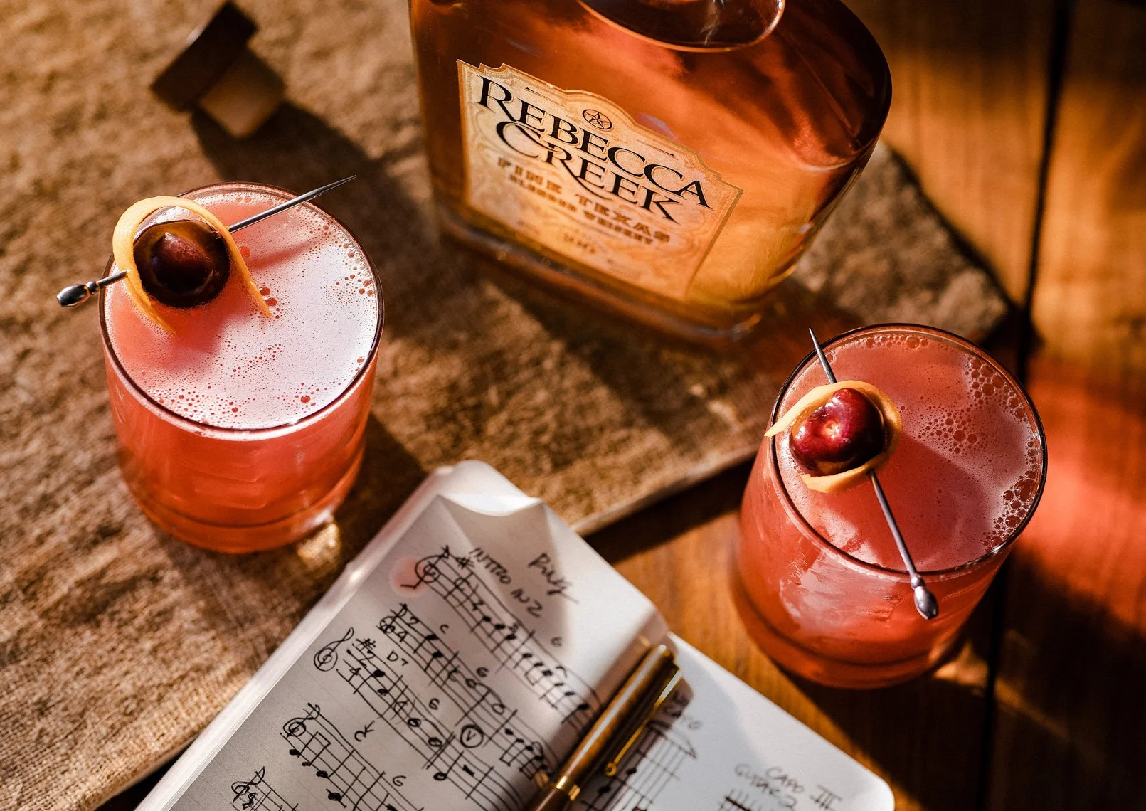 Two pink cocktails garnished with cherry and lemon peel, an open sheet of music, a pen, and a bottle labeled 'Rebbecca Creek' on a wooden table.