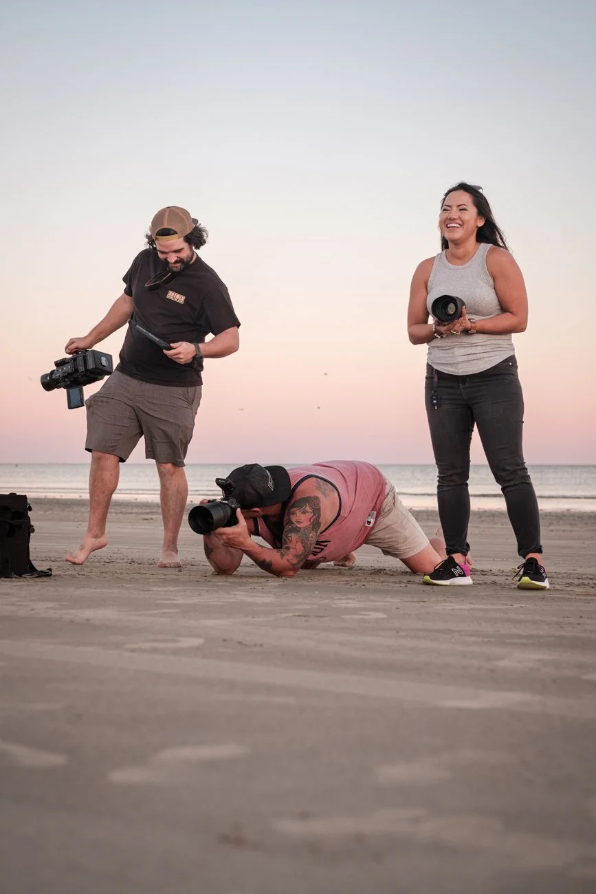 Three photographers on a beach during sunset, with one lying on the sand taking pictures, and the other two standing, holding camera equipment, smiling.