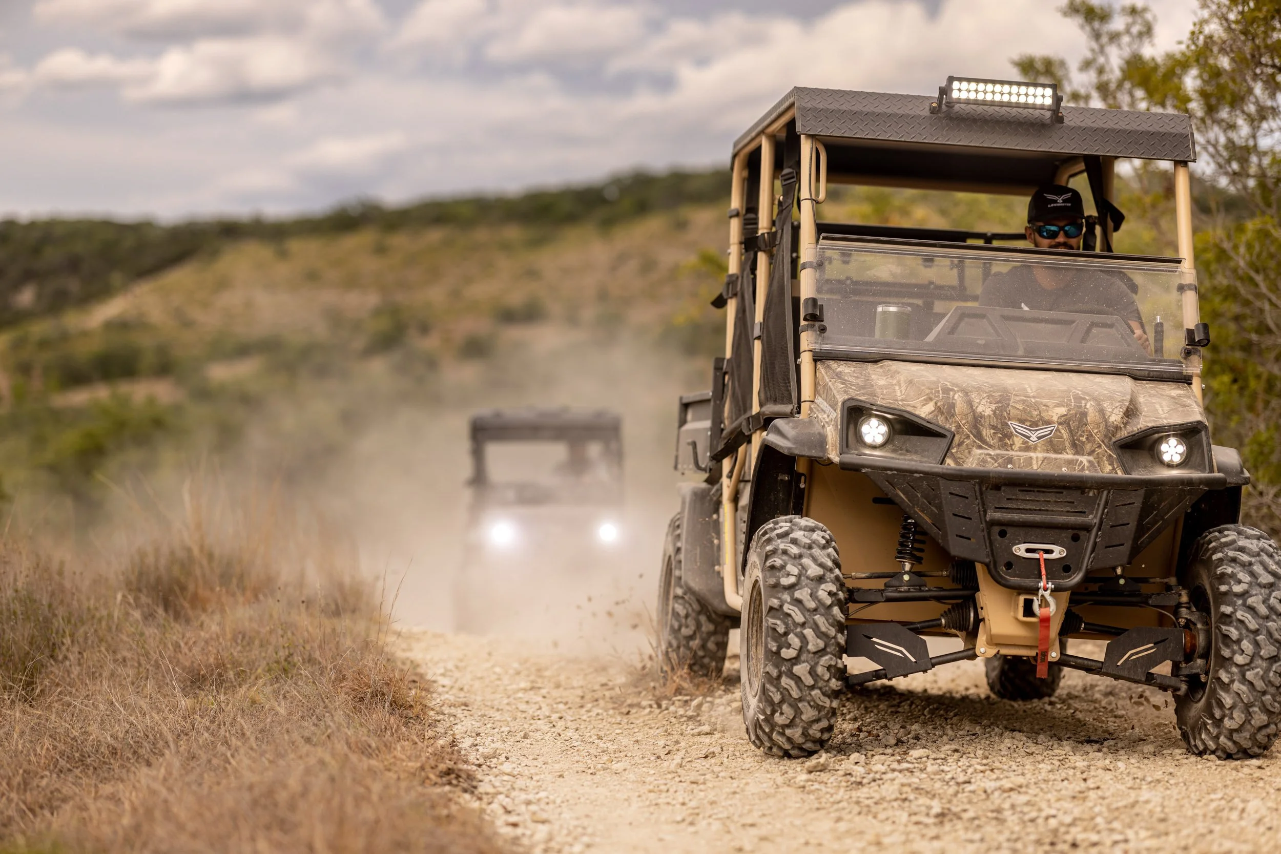 A person riding a camouflage all-terrain vehicle on a dirt trail in a rural outdoor setting with trees and a cloudy sky.