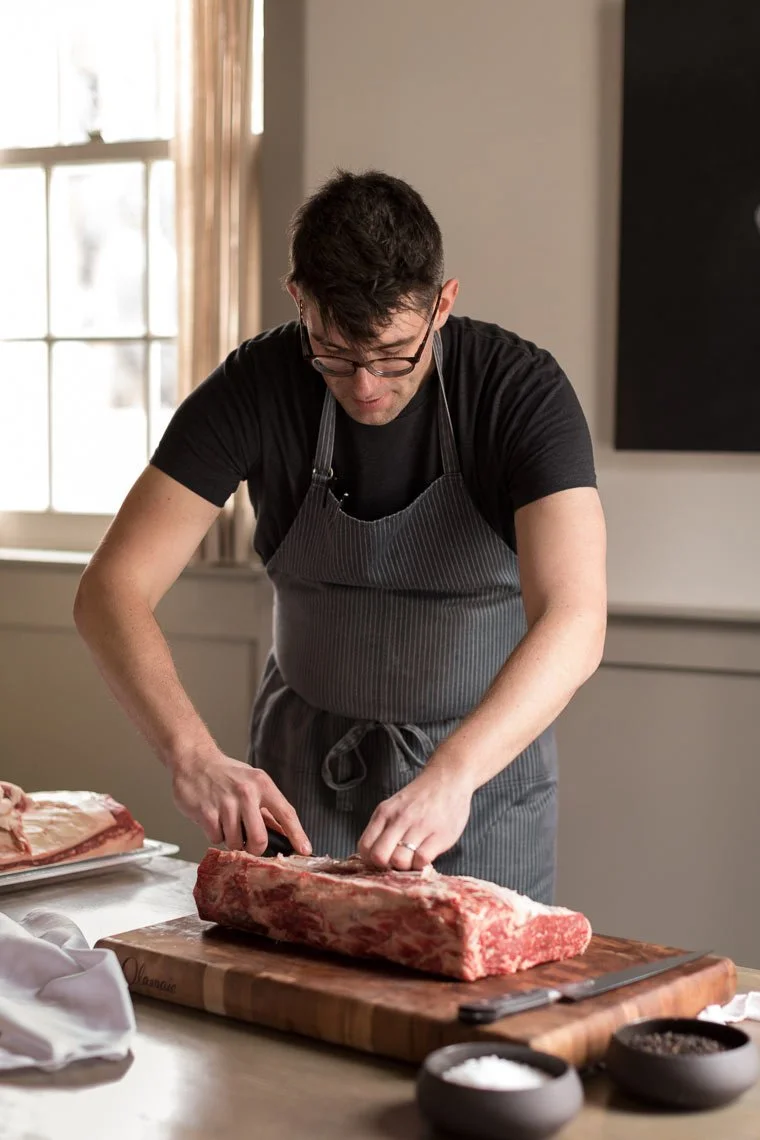 Man wearing glasses and a black shirt, with an apron, prepares a large piece of meat on a cutting board in a well-lit kitchen.