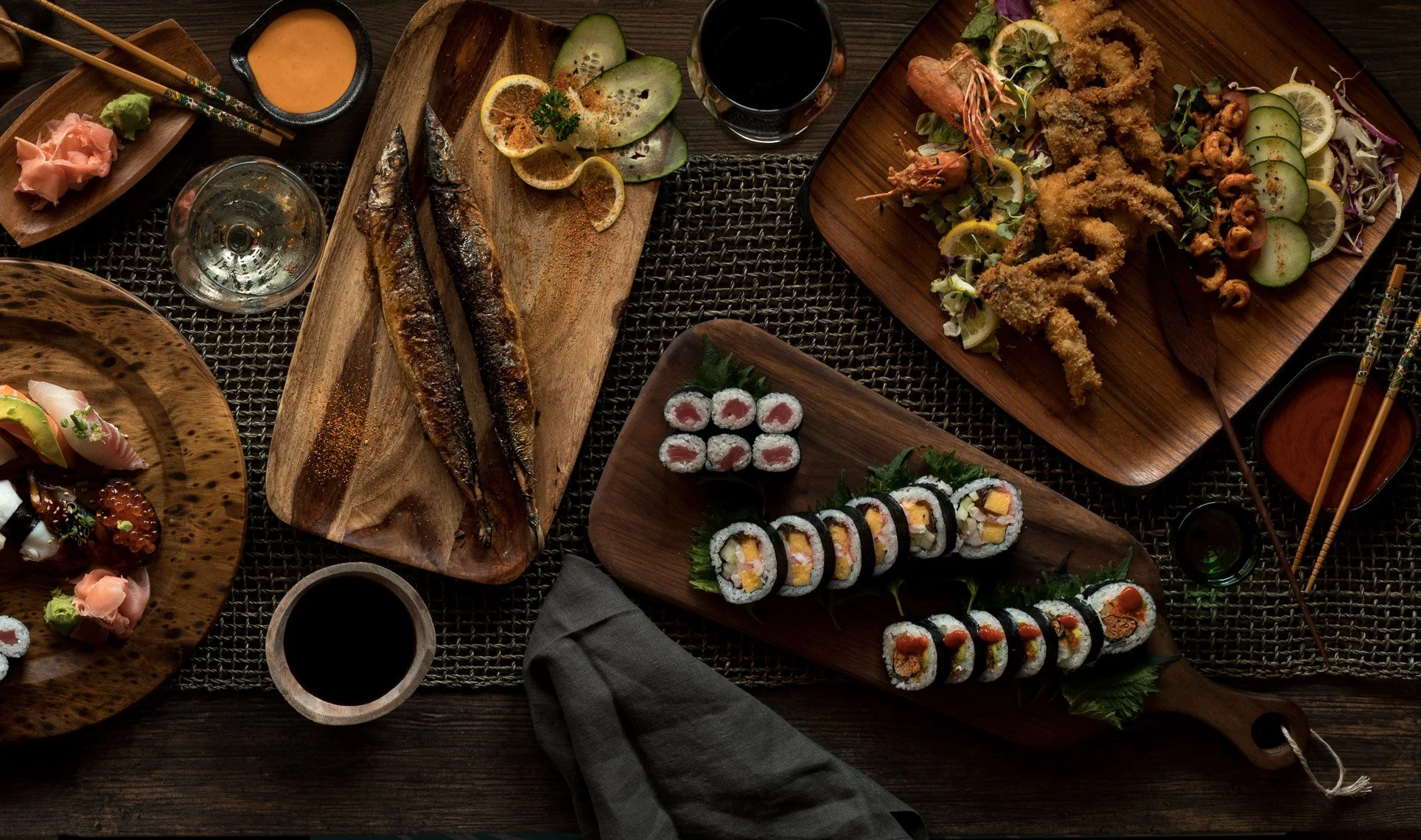 A variety of Japanese cuisine on wooden trays and plates, including sushi rolls, grilled fish, fried tempura, sashimi, and dipping sauces, arranged on a dark table.