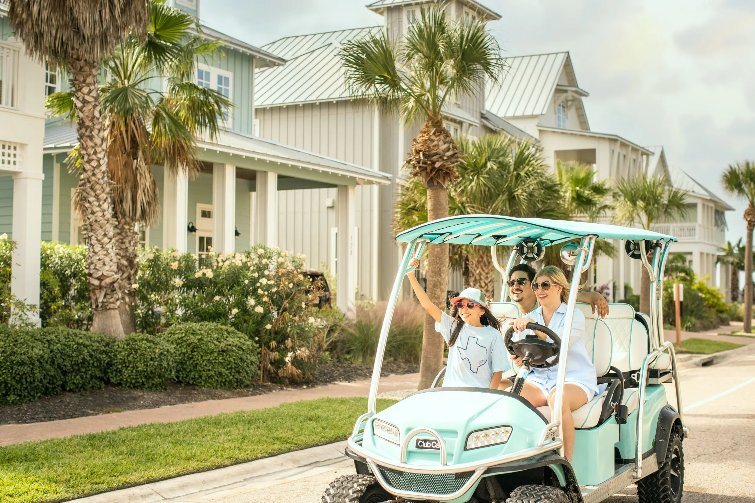 A family of four riding in a golf cart along a suburban street with palm trees and colorful houses in the background. The family is smiling and enjoying their ride.