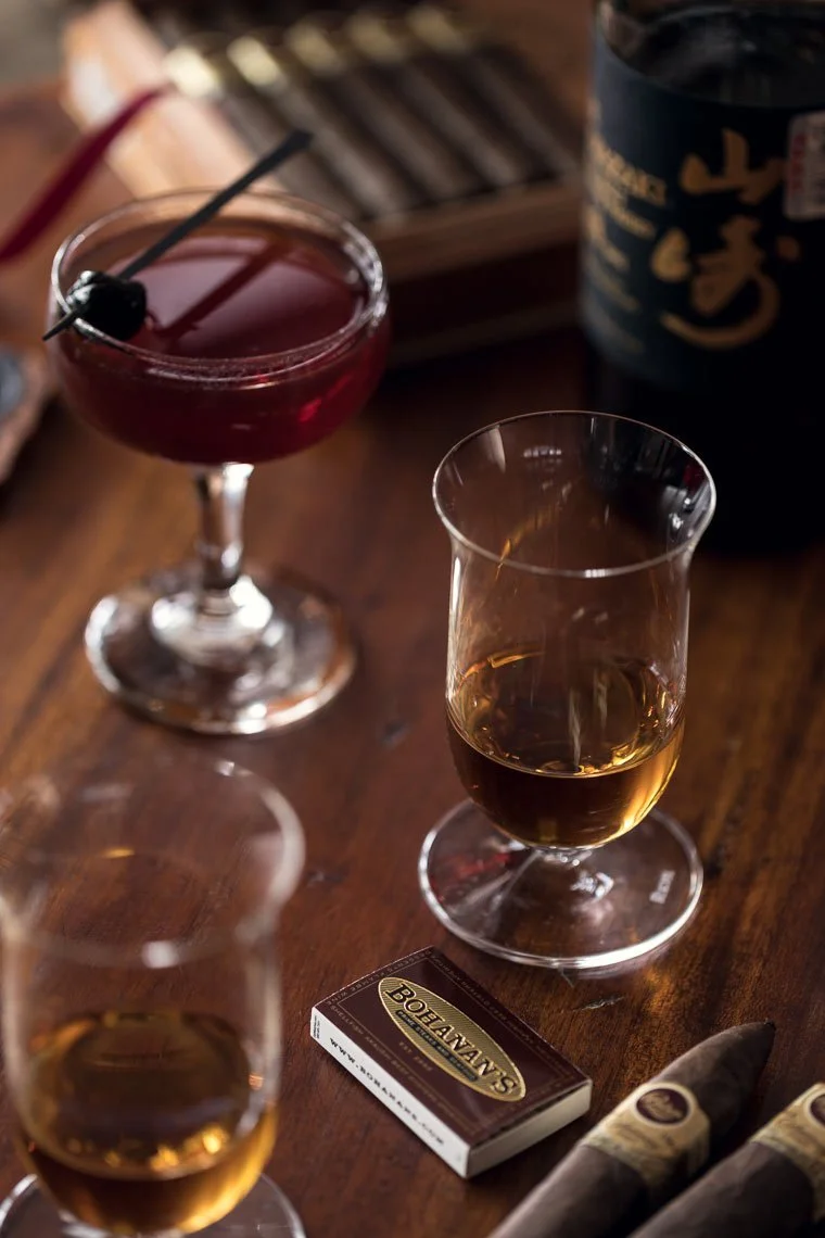 Two glasses of whiskey and one glass of red cocktail with cherry garnish on a wooden table, along with a pack of Bodegas cigarillos and two cigars.