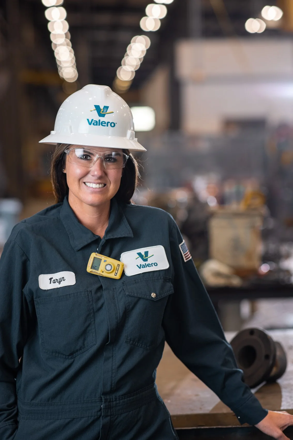 A woman wearing a dark work uniform, safety glasses, a white Hard hat with the 'Valero' logo, and a name tag that says 'Taryn' poses in a factory or workshop environment.