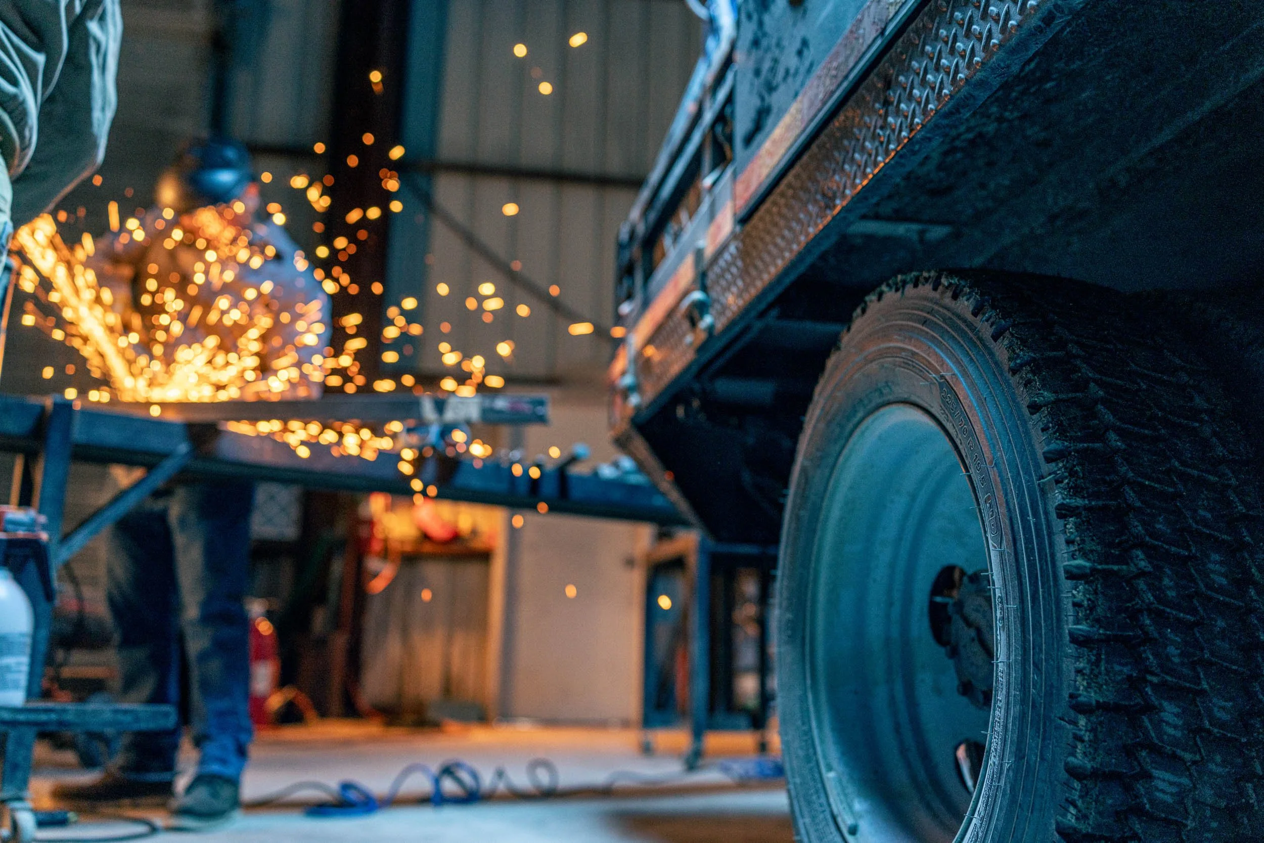 Person welding metal on a workbench in a workshop, with sparks flying, a truck tire visible in the foreground, and tools and equipment in the background.