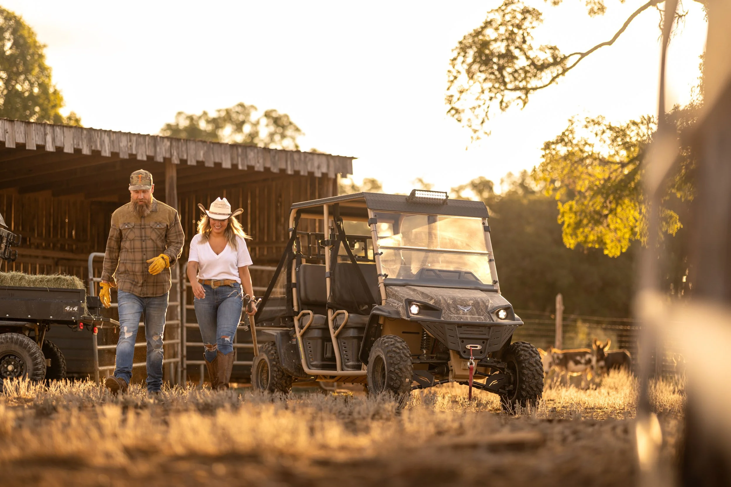 A man and woman walking together outdoors near farm equipment, a golf cart, and a wooden barn, with trees and sunset in the background.