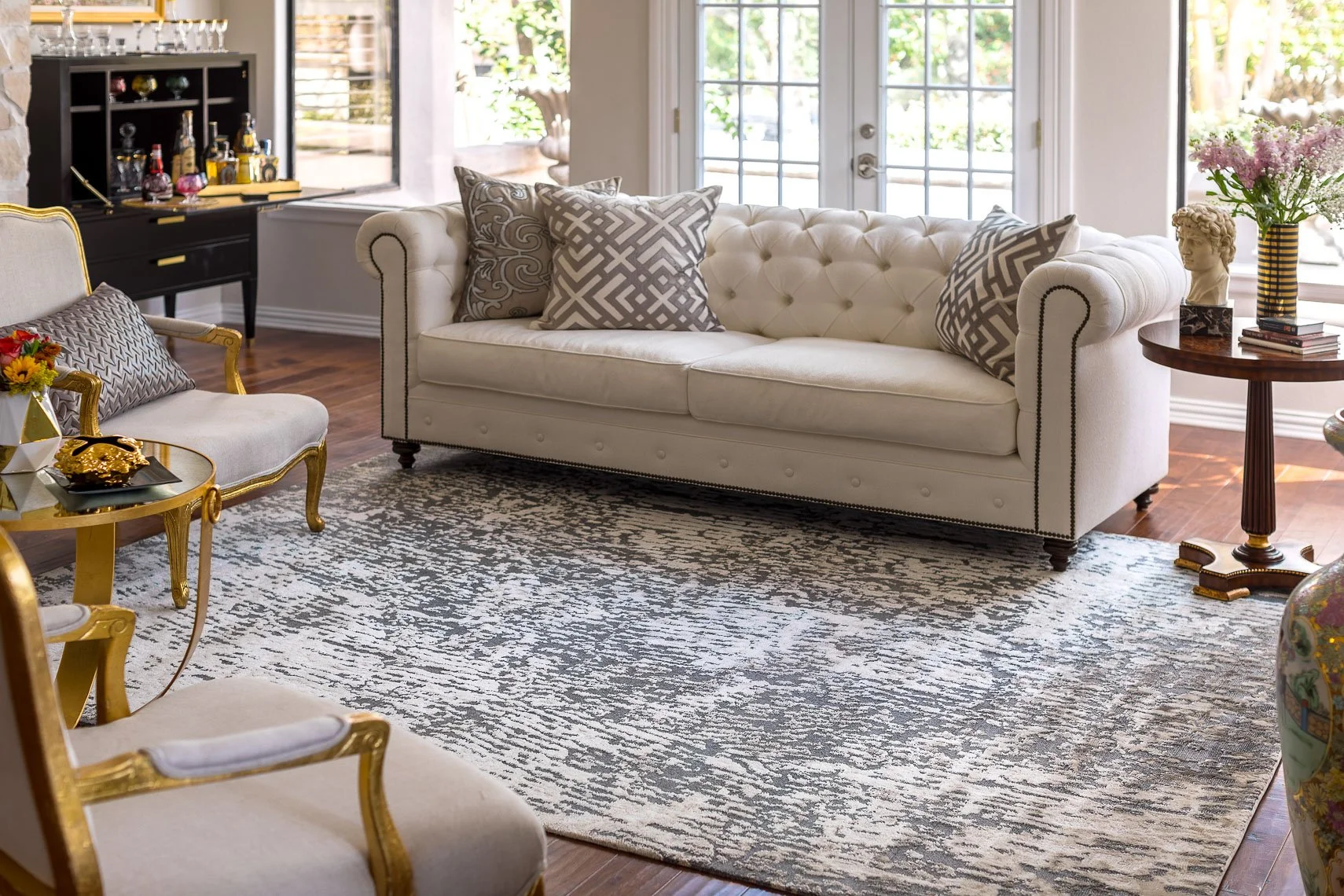 Living room with white tufted sofa, decorative pillows, a side table with flowers, a patterned rug, and a glass door leading outside.