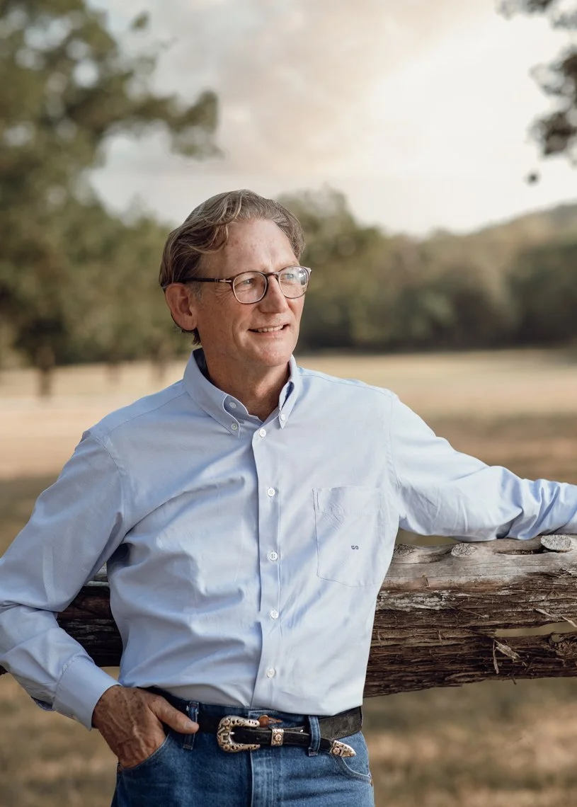 A middle-aged man wearing glasses and a light blue button-down shirt standing outdoors near a log with a field and trees in the background on a cloudy day.