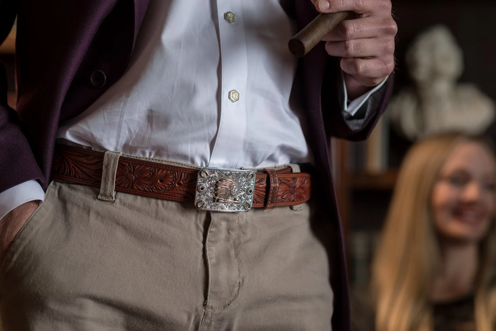Close-up of a man wearing a white button-up shirt, a maroon blazer, a brown leather belt with an ornate silver buckle, khaki pants, holding a cigar, with a woman smiling in the background.