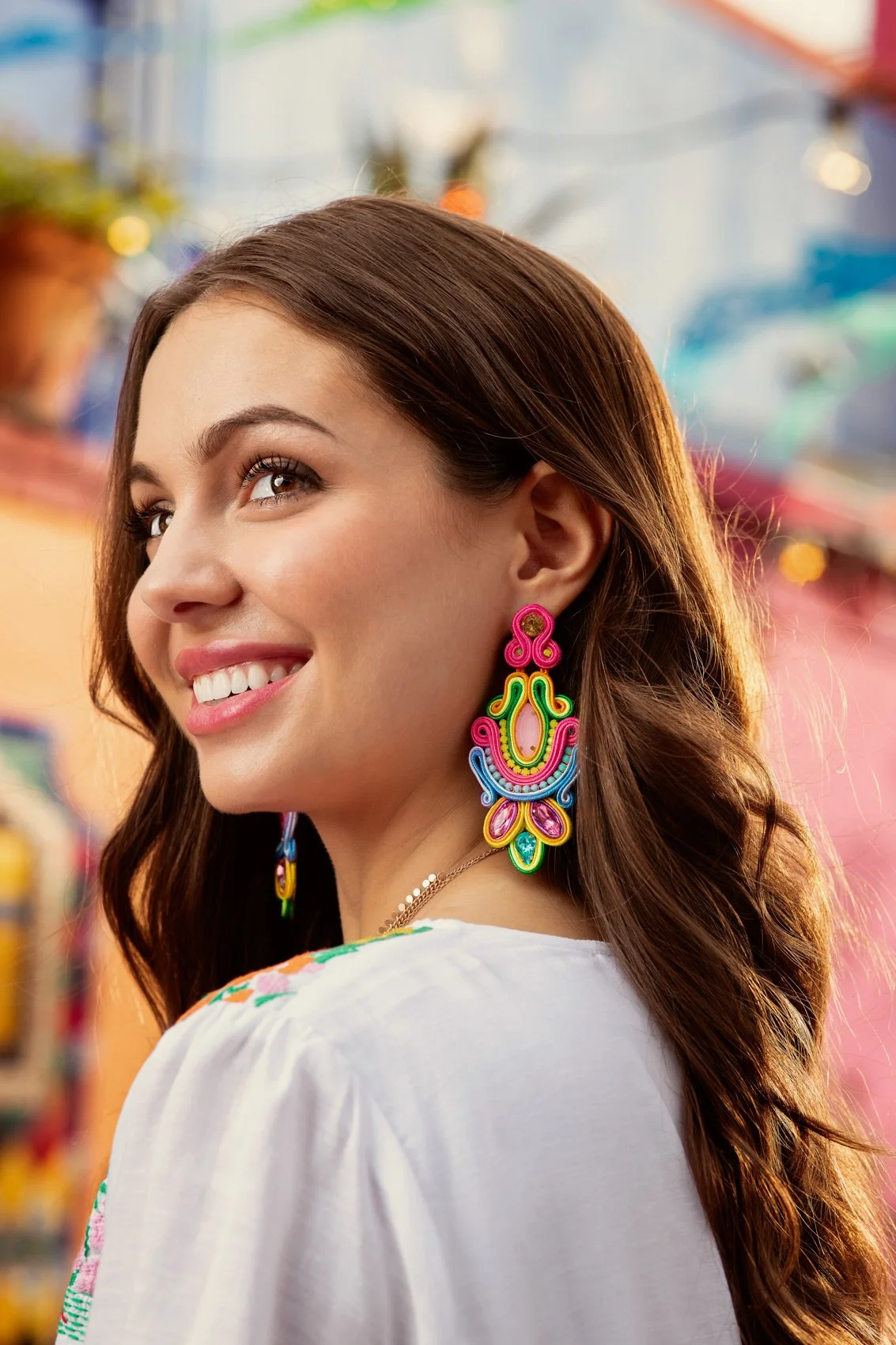 Close-up of a smiling woman with long brown hair wearing large, colorful, decorative earrings in a vibrant market or store with blurred background of various items and colorful lighting.