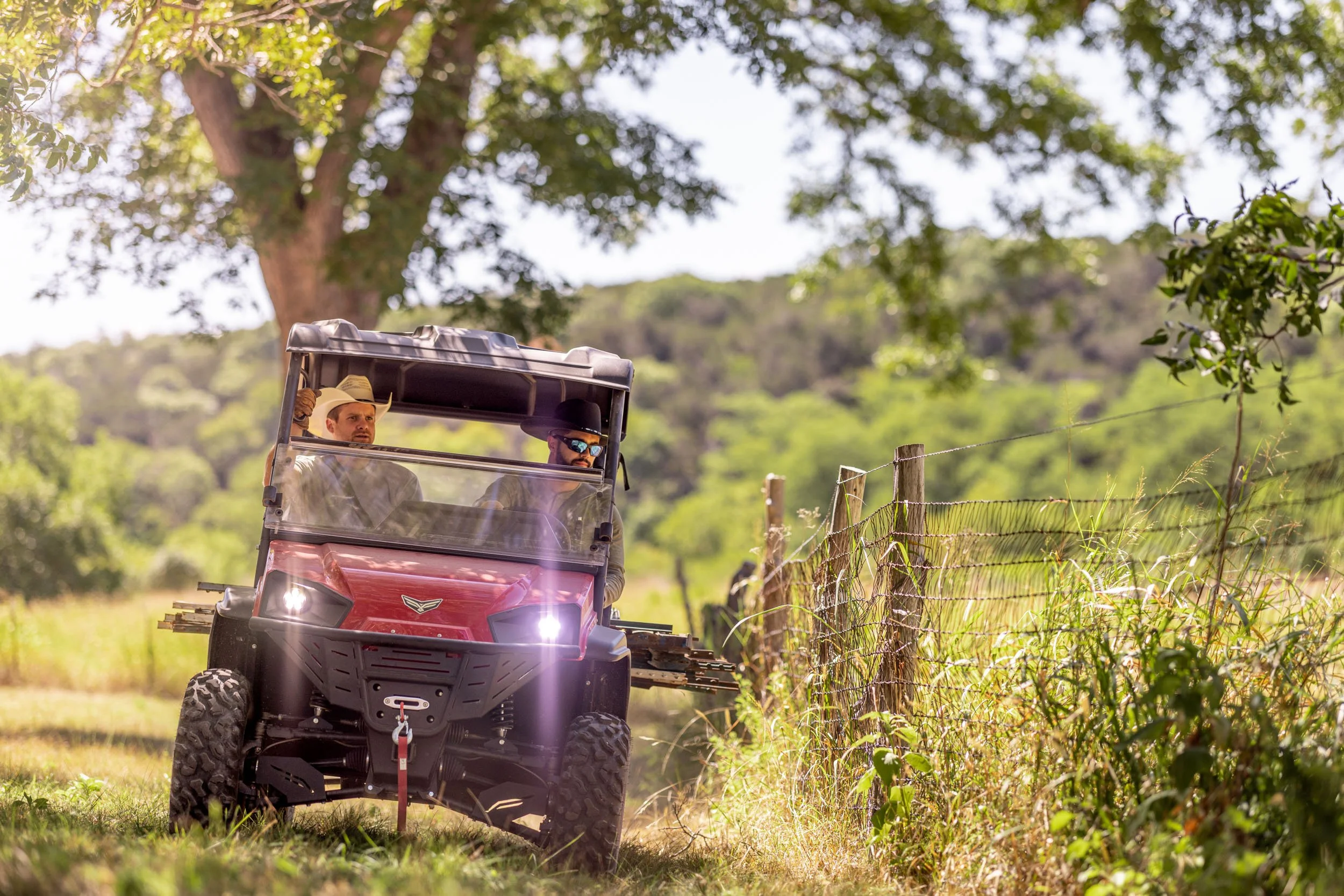 Two men riding a red utility vehicle along a fence in a rural area with trees and green hills in the background.