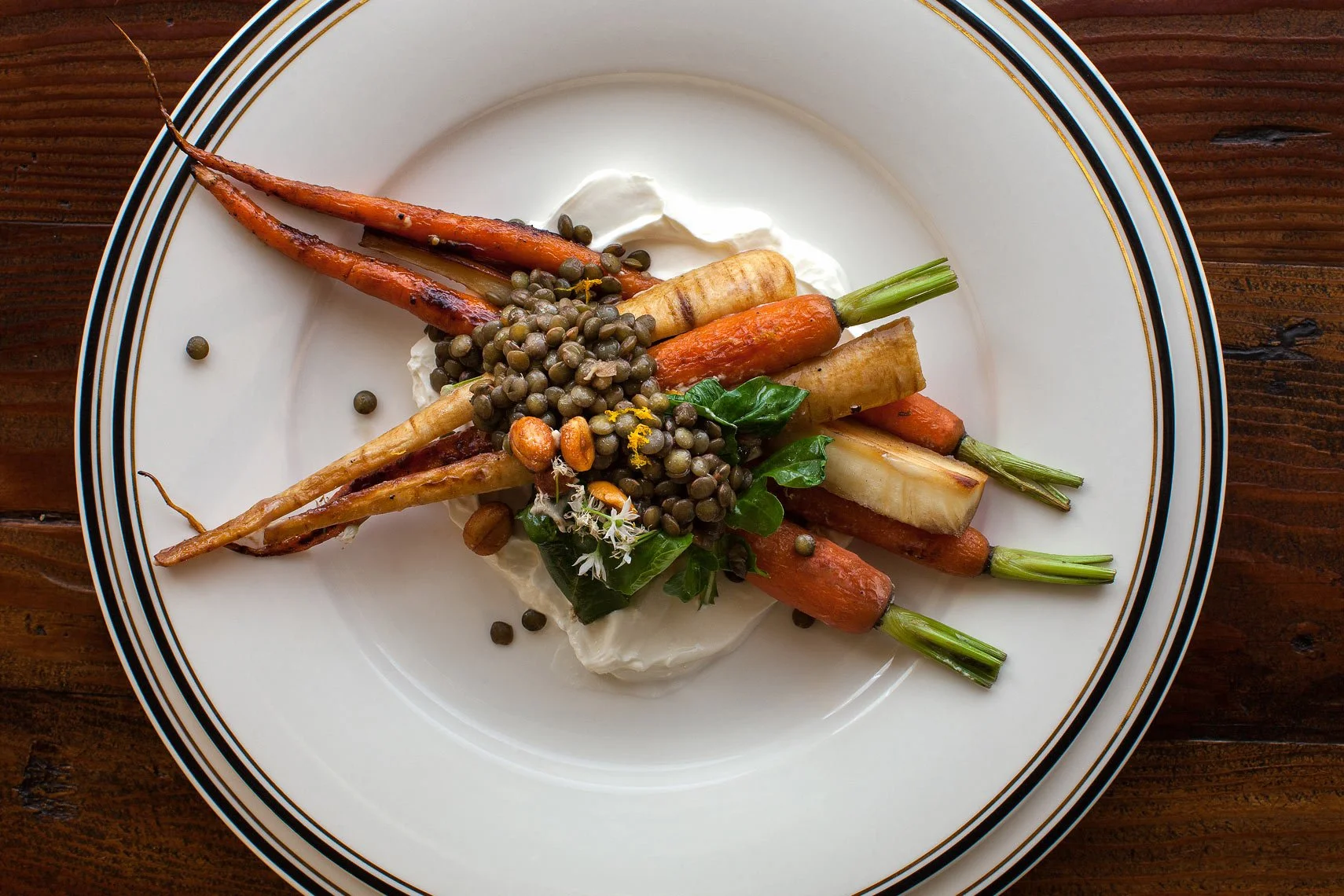 Roasted carrots, parsnips, and green onions garnished with black lentils, chickpeas, and fresh herbs on a white plate with gold and black rim, on a wooden surface.