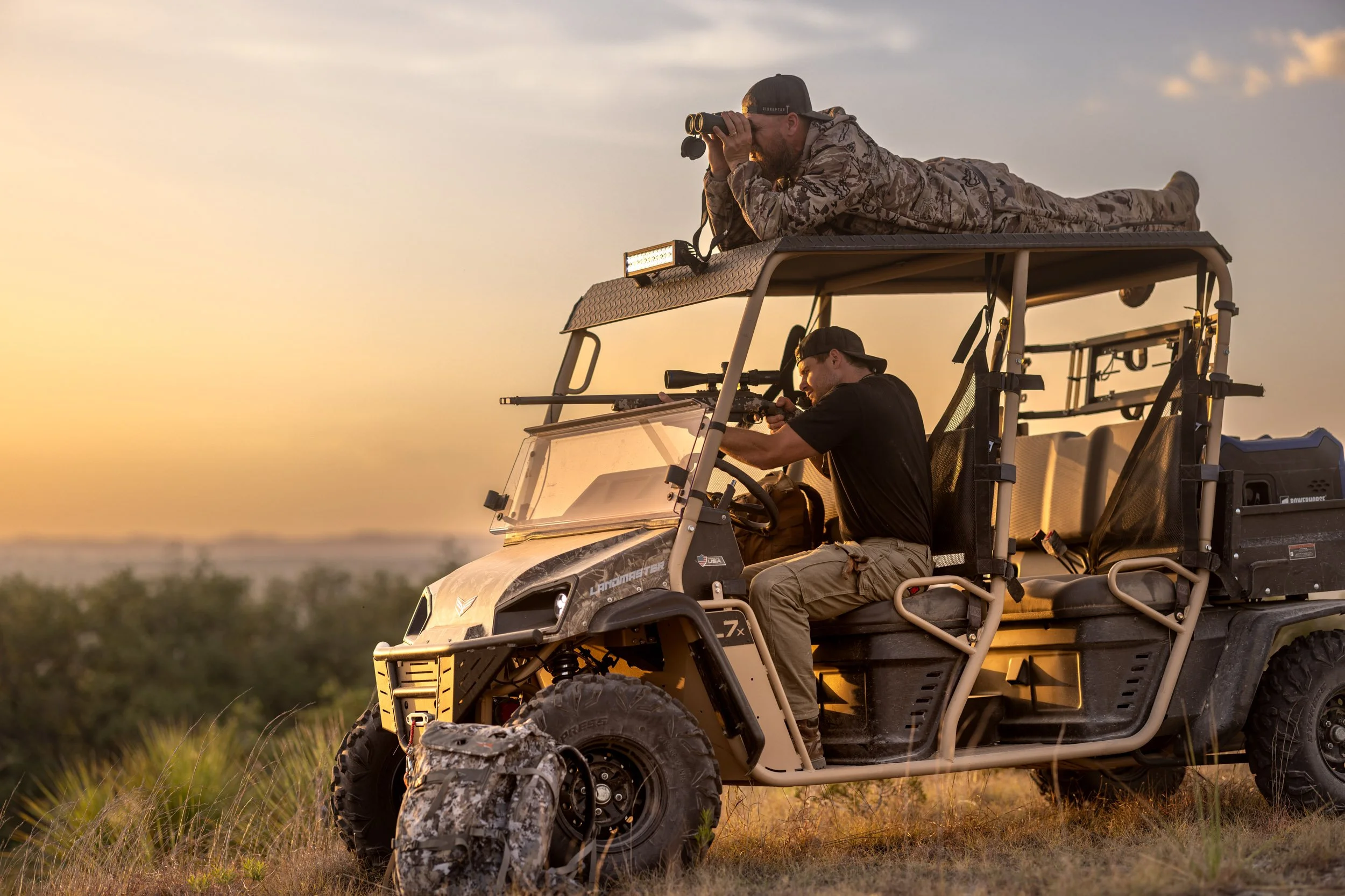 Two men are on a beige off-road utility vehicle in a grassland area during sunset. One man is lying on top of the vehicle with binoculars, looking into the distance, wearing camouflage clothing and a cap. The other man is sitting inside the vehicle, 