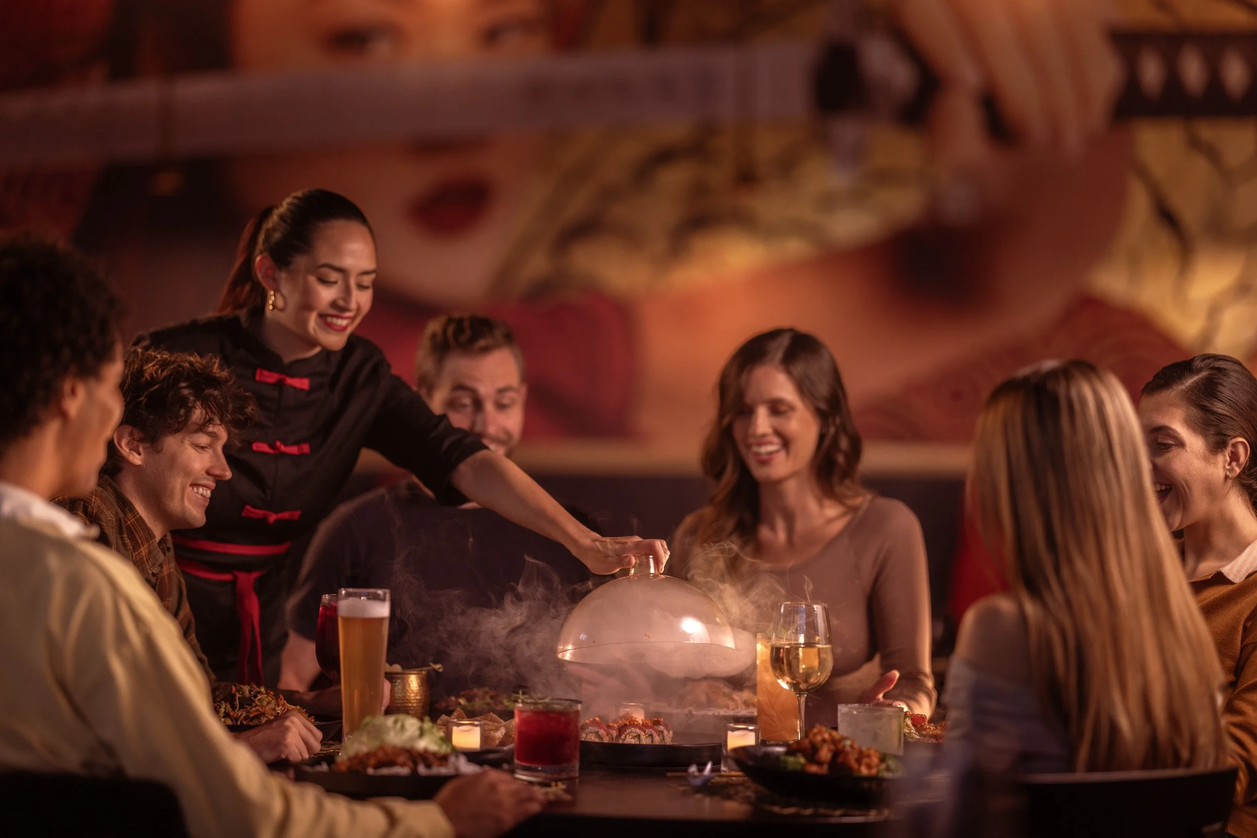 A group of people sitting around a dinner table with food and drinks, while a woman in a black dress with red accents serves the meal in a warmly lit restaurant. There are several beverages including wine and beer, and small candles on the table.
