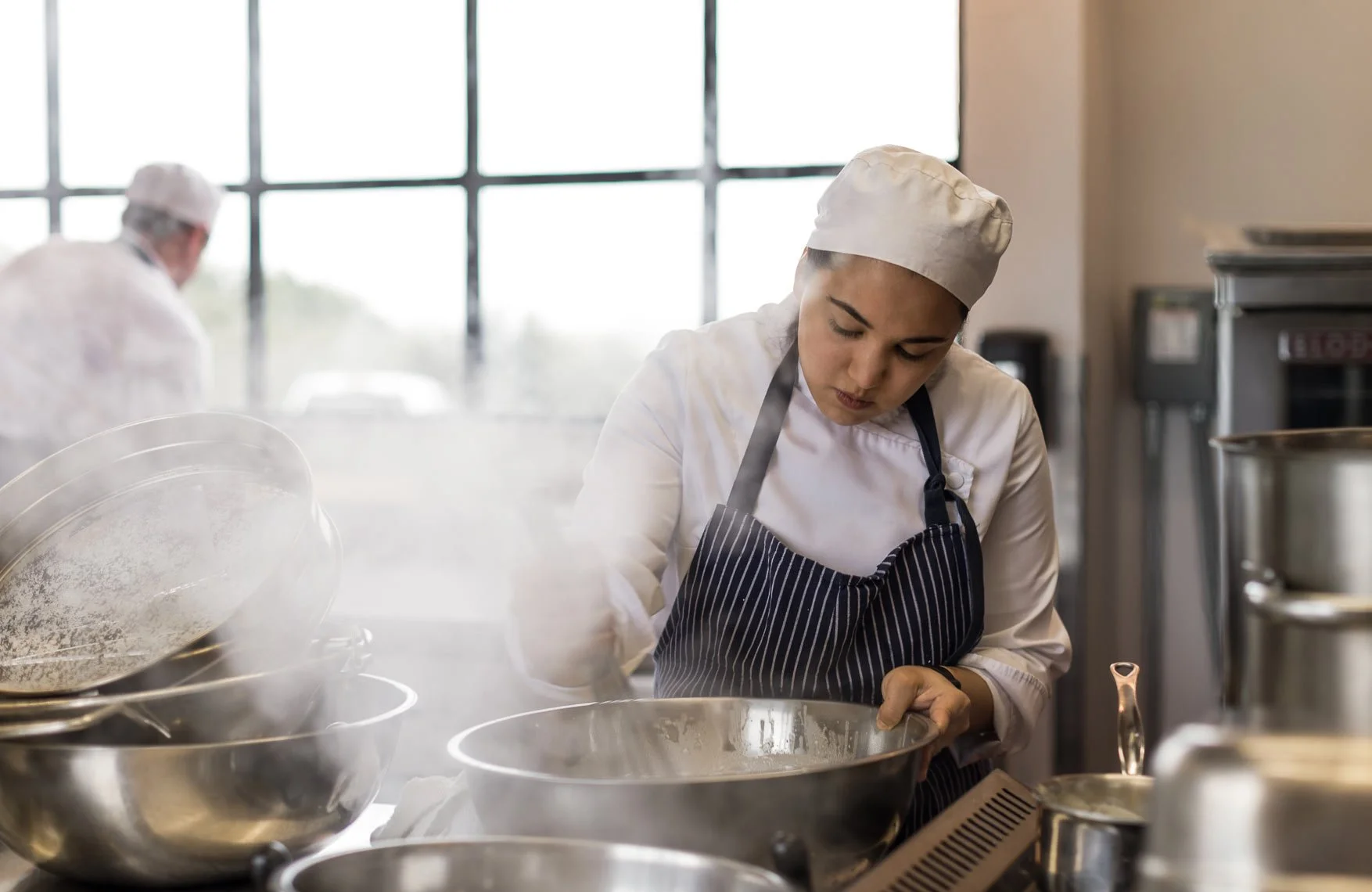 A female chef in a white uniform and striped apron, wearing a chef hat, focusing on her work in a commercial kitchen with steam and stainless steel pots.