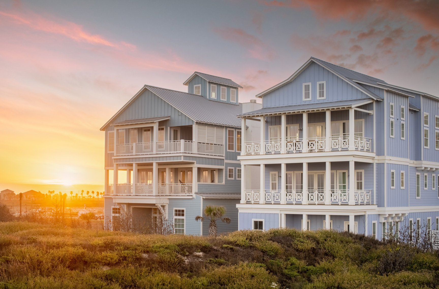A blue multi-story beach house with multiple balconies during sunset, surrounded by greenery and palm trees.