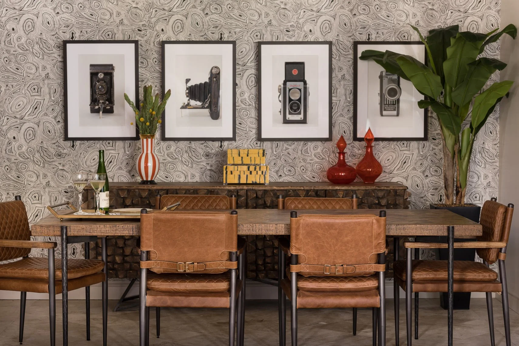 A dining room with a large wooden table and six brown leather chairs. The wall has black-and-white framed photographs of vintage cameras. There are decorative items on the sideboard, including a tall orange and white striped vase with flowers, a ster