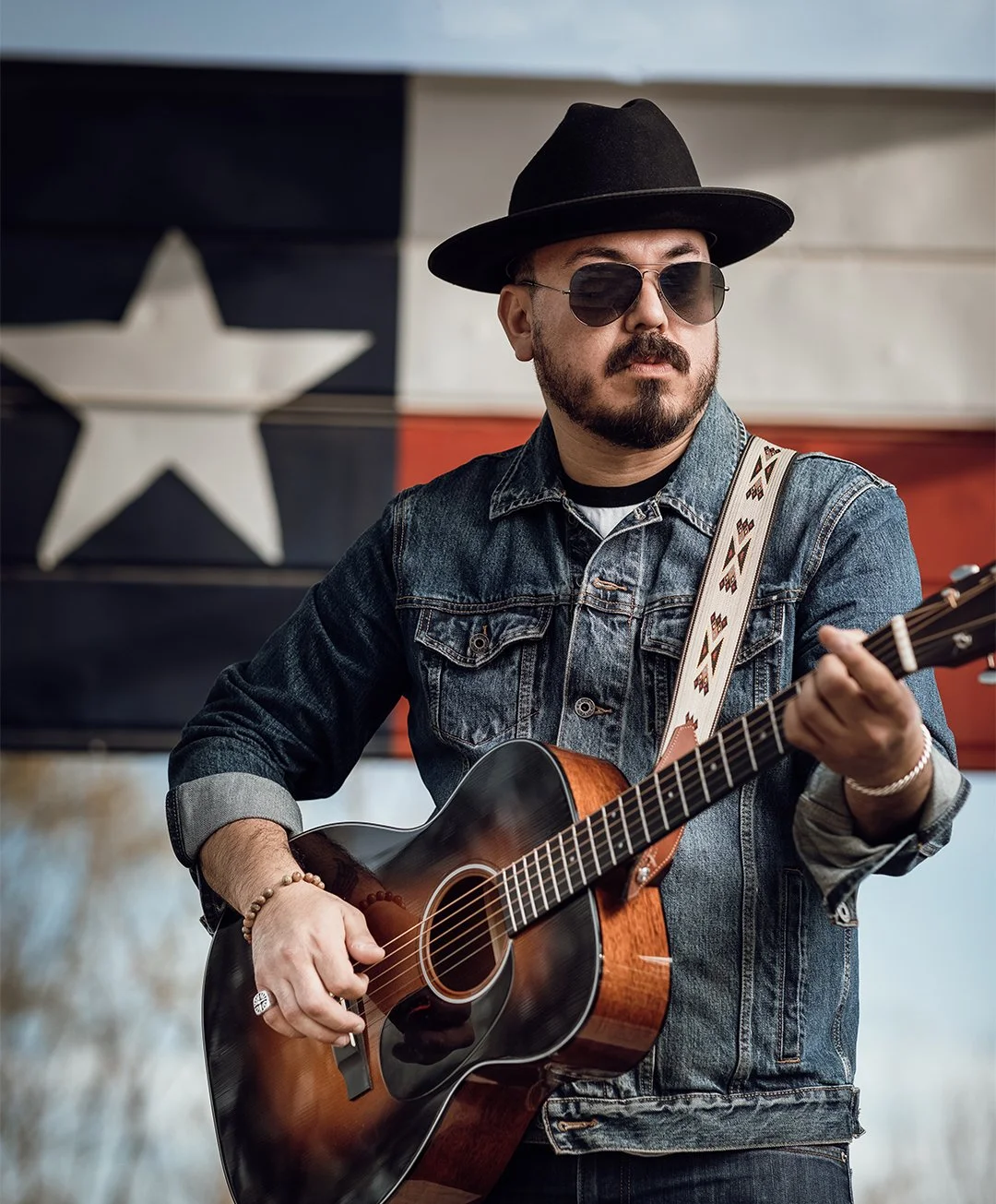 A man wearing sunglasses, a black hat, and a denim jacket plays an acoustic guitar in front of a Texas flag.