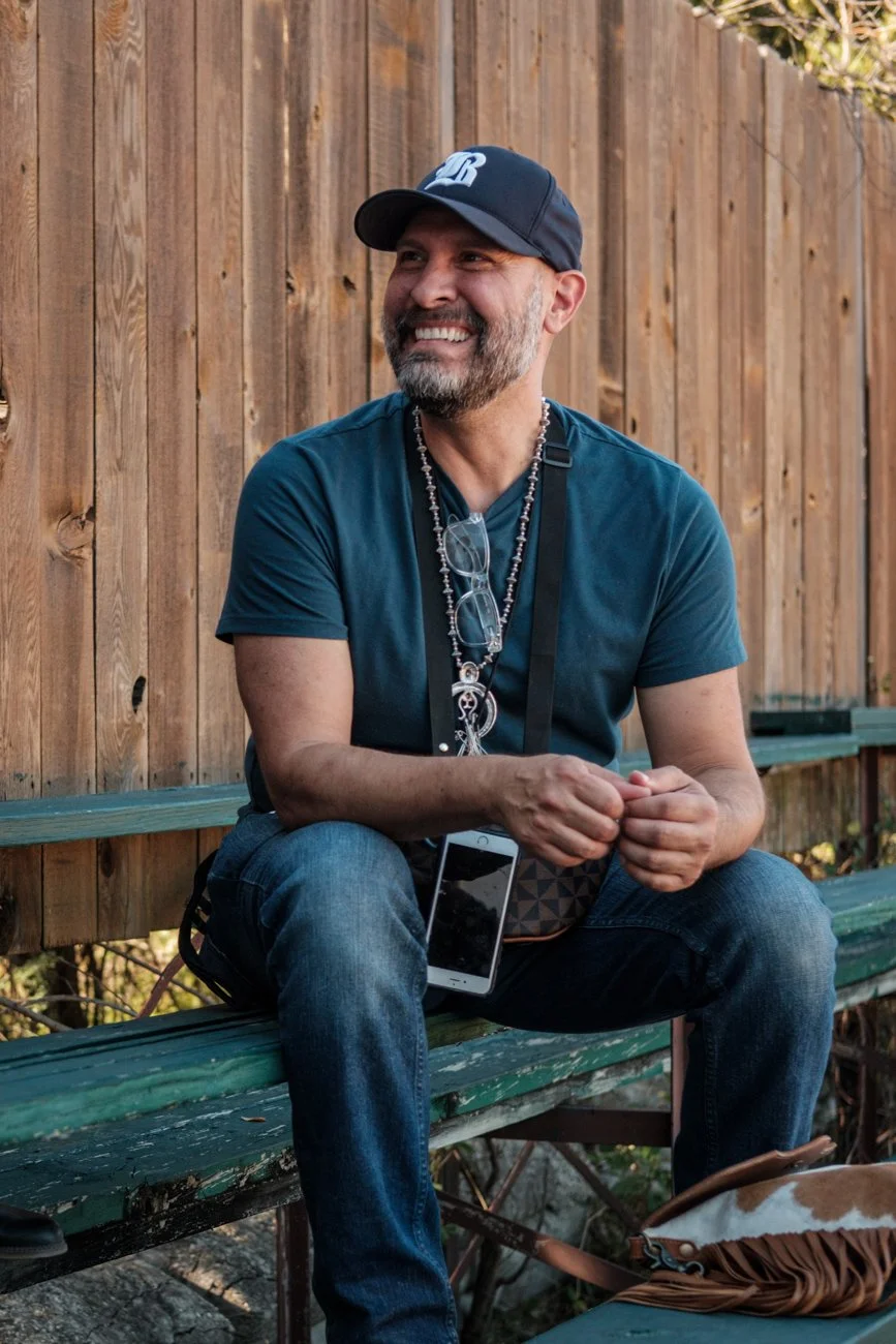 A smiling man with a beard and glasses hanging from a necklace sitting on a green park bench in front of a wooden fence, wearing a dark blue T-shirt, jeans, and a black baseball cap, holding a smartphone and clasping his hands.