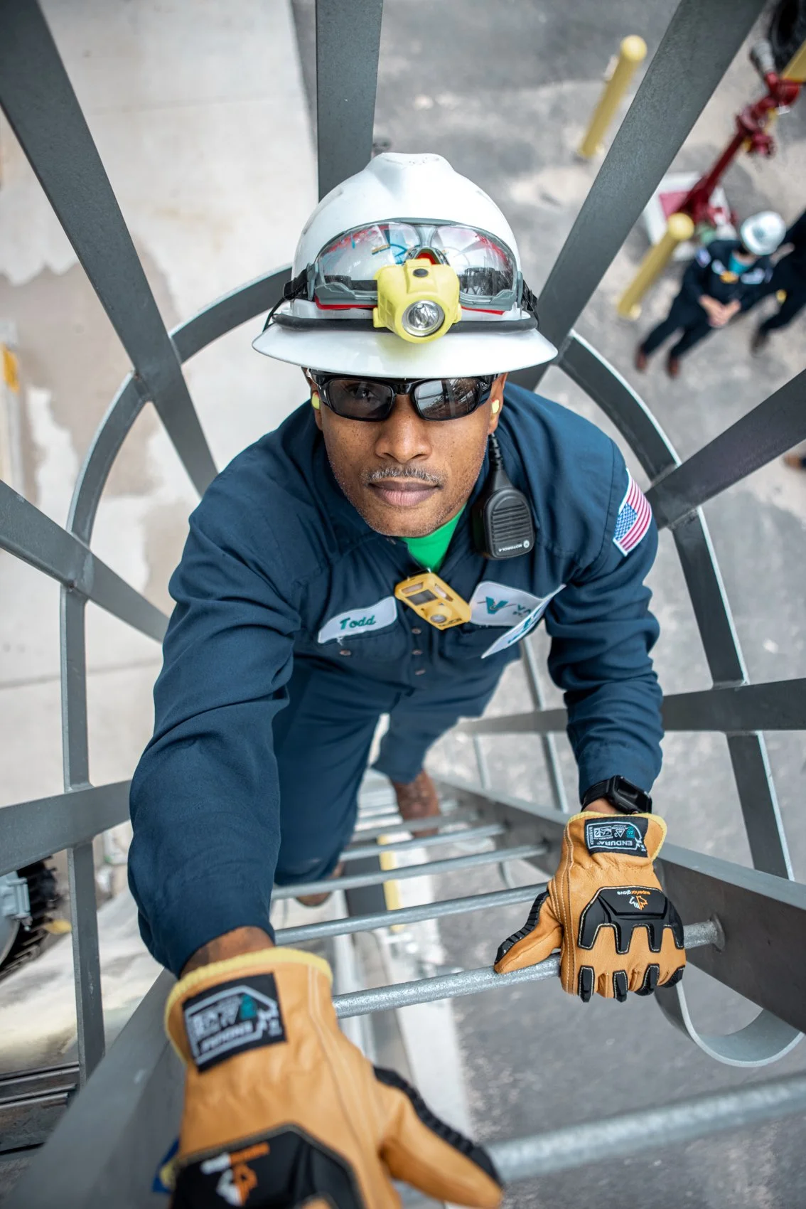 A firefighter wearing a helmet with a headlamp and sunglasses climbing a metal ladder, with safety gloves and a walkie-talkie clipped to his uniform, at a construction site or emergency scene.