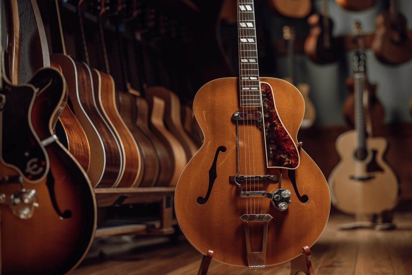 A vintage hollow-body electric guitar with a sunburst finish and tortoiseshell pickguard, standing on a guitar stand in a music store or guitar shop, surrounded by other acoustics and electrics on display.