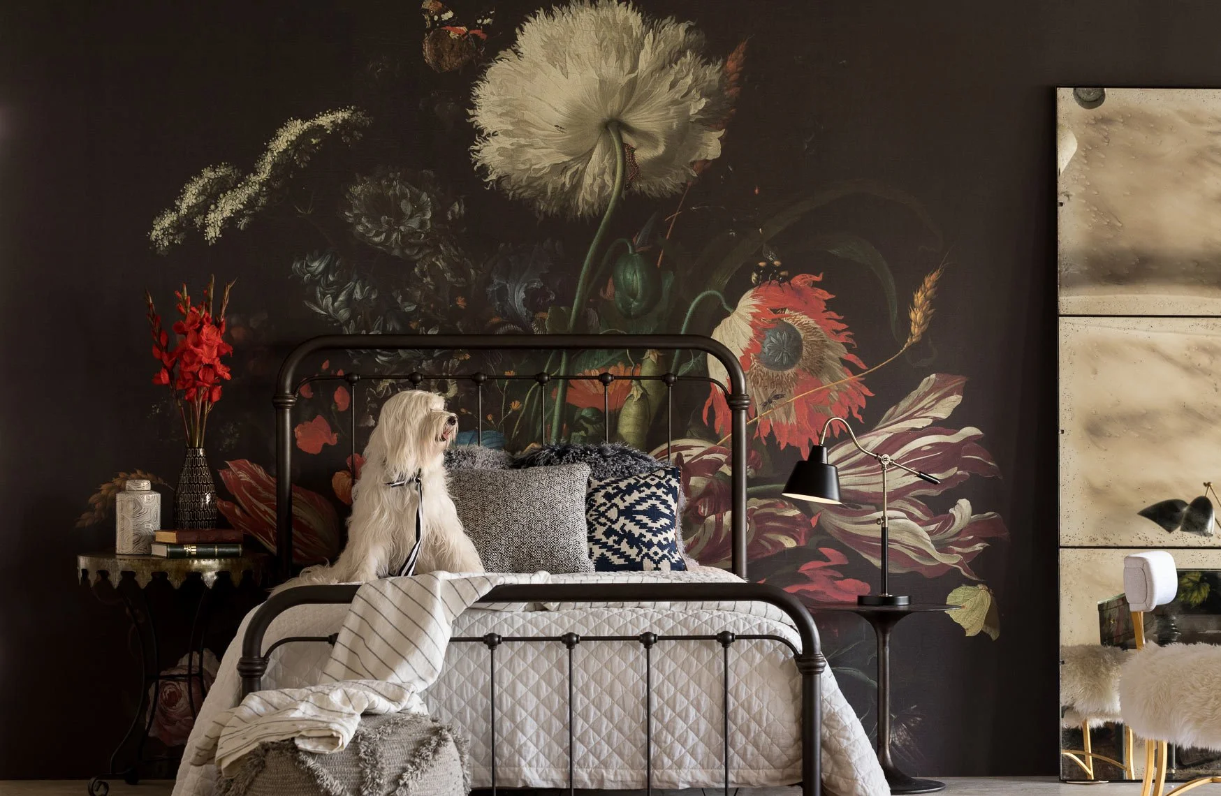 A bedroom with a dark floral accent wall, a black metal bed frame, and a white dog sitting on the bed. The bed has pillows in various patterns and a blanket. To the left, there is a side table with a vase of red flowers and decorative jars. On the ri
