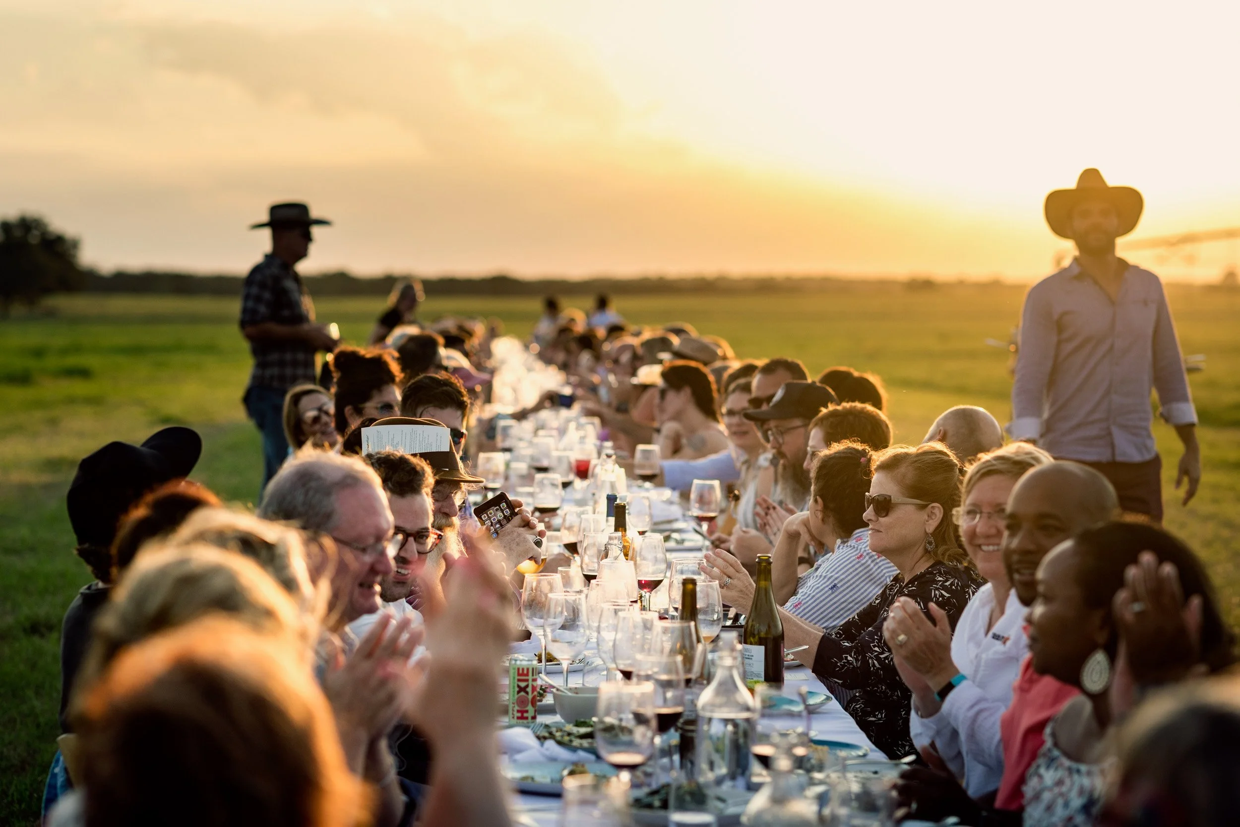 A large group of people gathered for an outdoor dinner at sunset, sitting at a long table with drinks and food, in a green field.