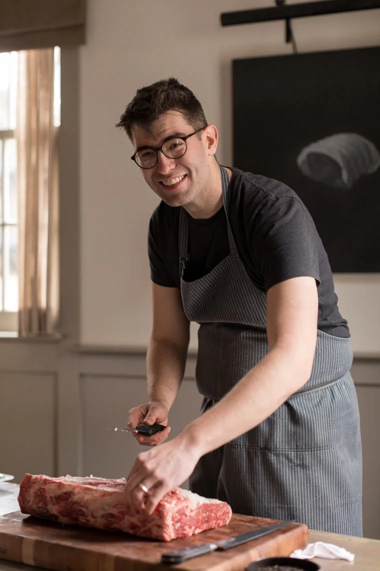 A man in glasses and a striped apron slicing a large piece of meat on a wooden cutting board in a well-lit kitchen.