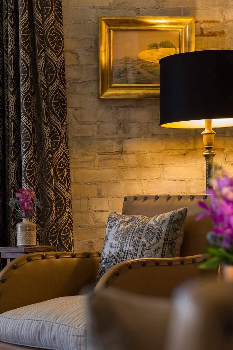 A cozy corner of a room with a leather armchair, patterned pillow, and a side table with flowers, a fringed curtain, a brass picture frame with a landscape, a black lampshade, and a brick wall.