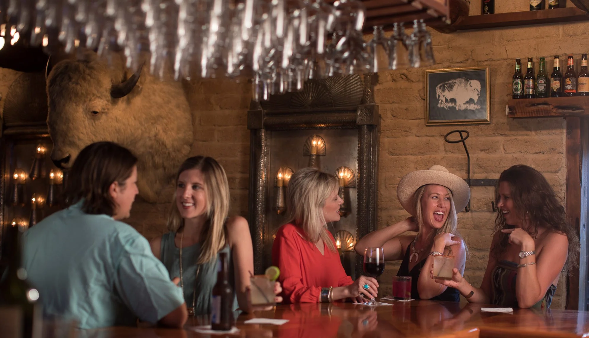 A group of women enjoying drinks and conversation at a bar with a rustic brick interior, wine glasses hanging overhead, a buffalo head mount, and framed artwork on the wall.