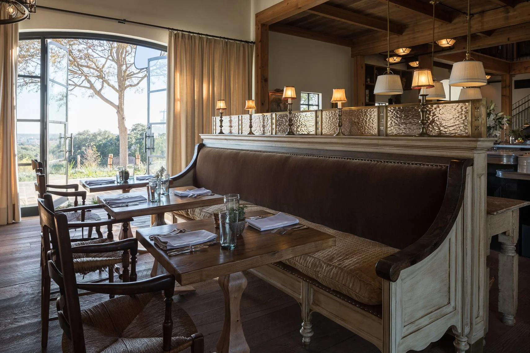 Interior of a cozy dining area with wooden furniture, a mix of chairs and a bench, set with napkins, glasses, and utensils, near large open windows with beige curtains revealing outdoor trees.
