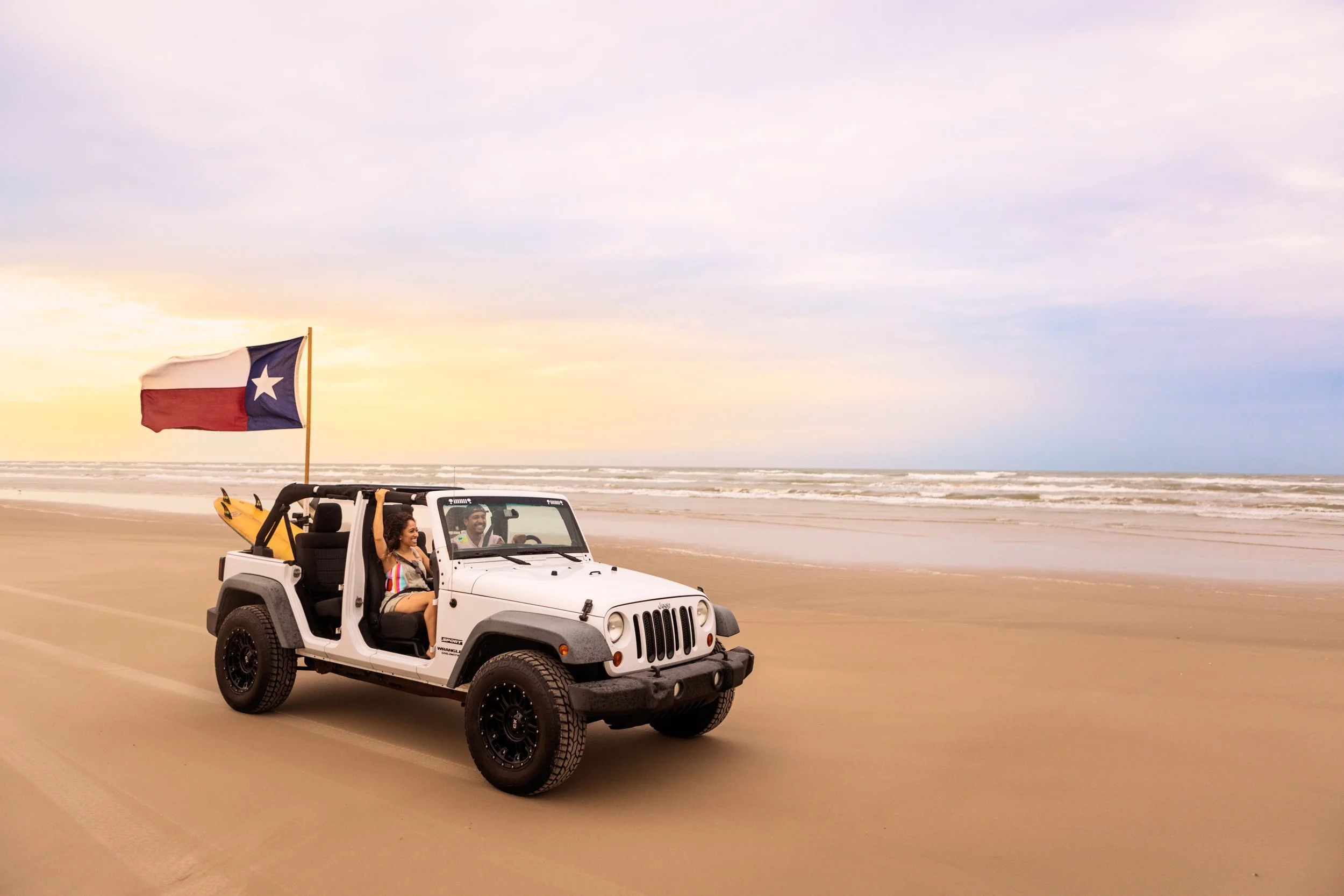 A white Jeep with two people in it on the beach. The woman is holding an American flag and the man is driving, both smiling. There are surfboards attached to the vehicle's back, and the beach stretches into the distance with the ocean and sky under a