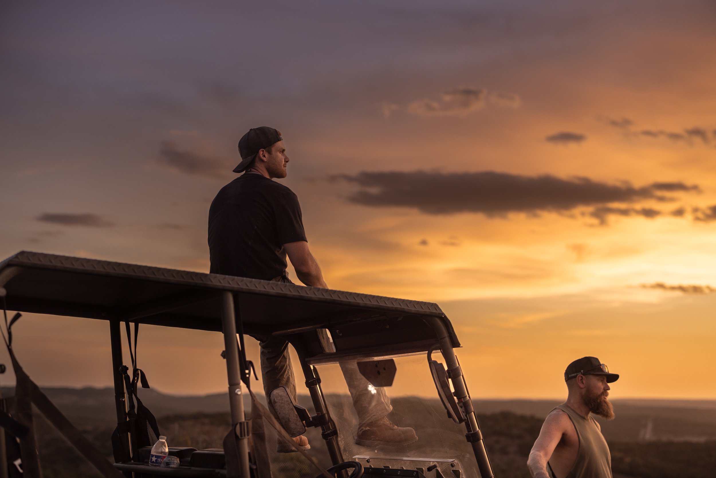 Two men sit on golf carts outdoors during sunset, one wearing a black cap and black t-shirt looking to the left, the other shirtless wearing glasses, both with a landscape in the background.