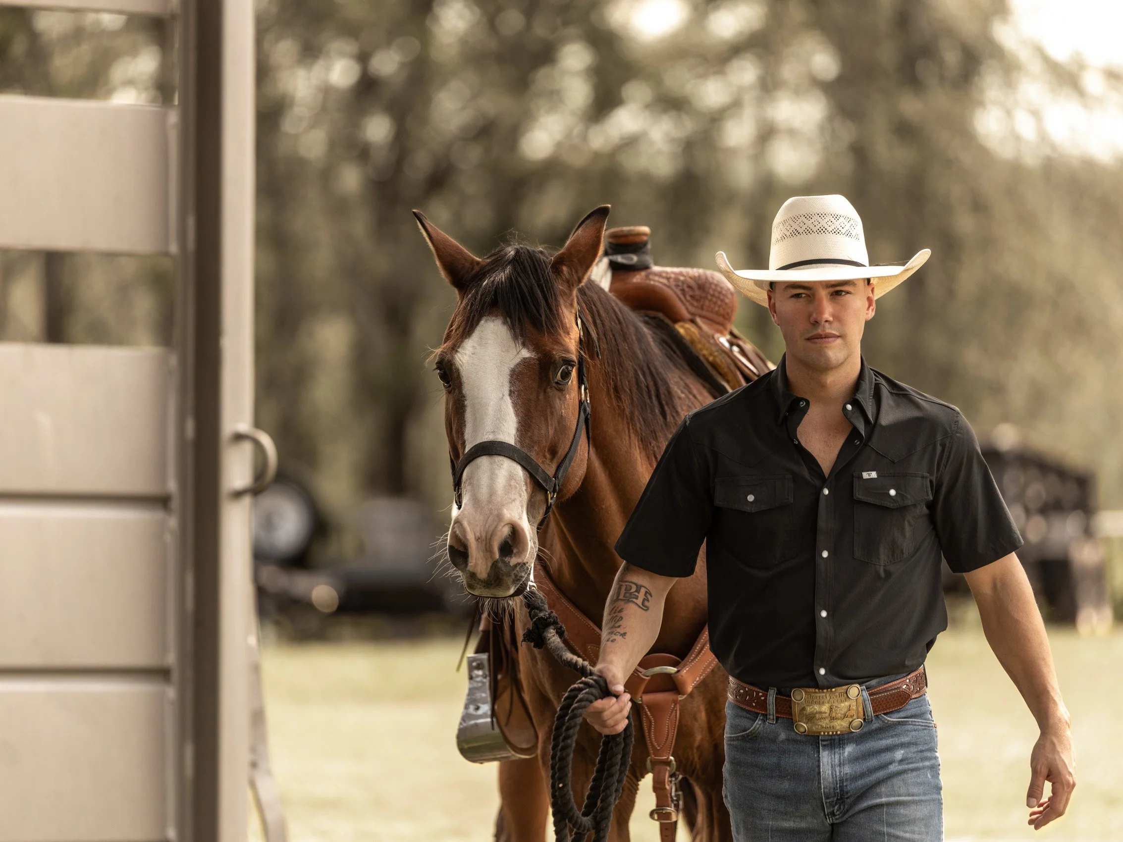 A man wearing a white cowboy hat and black shirt walks beside a brown and white horse on a trail in a wooded area.