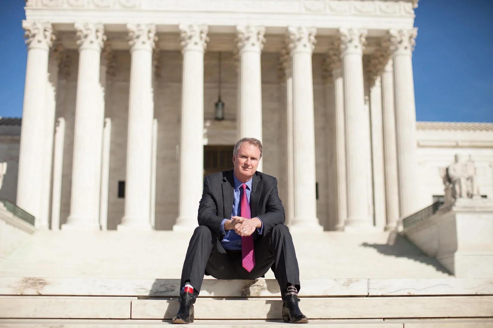 A man in a business suit sitting on steps in front of the U.S. Supreme Court building, with large white columns and classical architecture behind him.