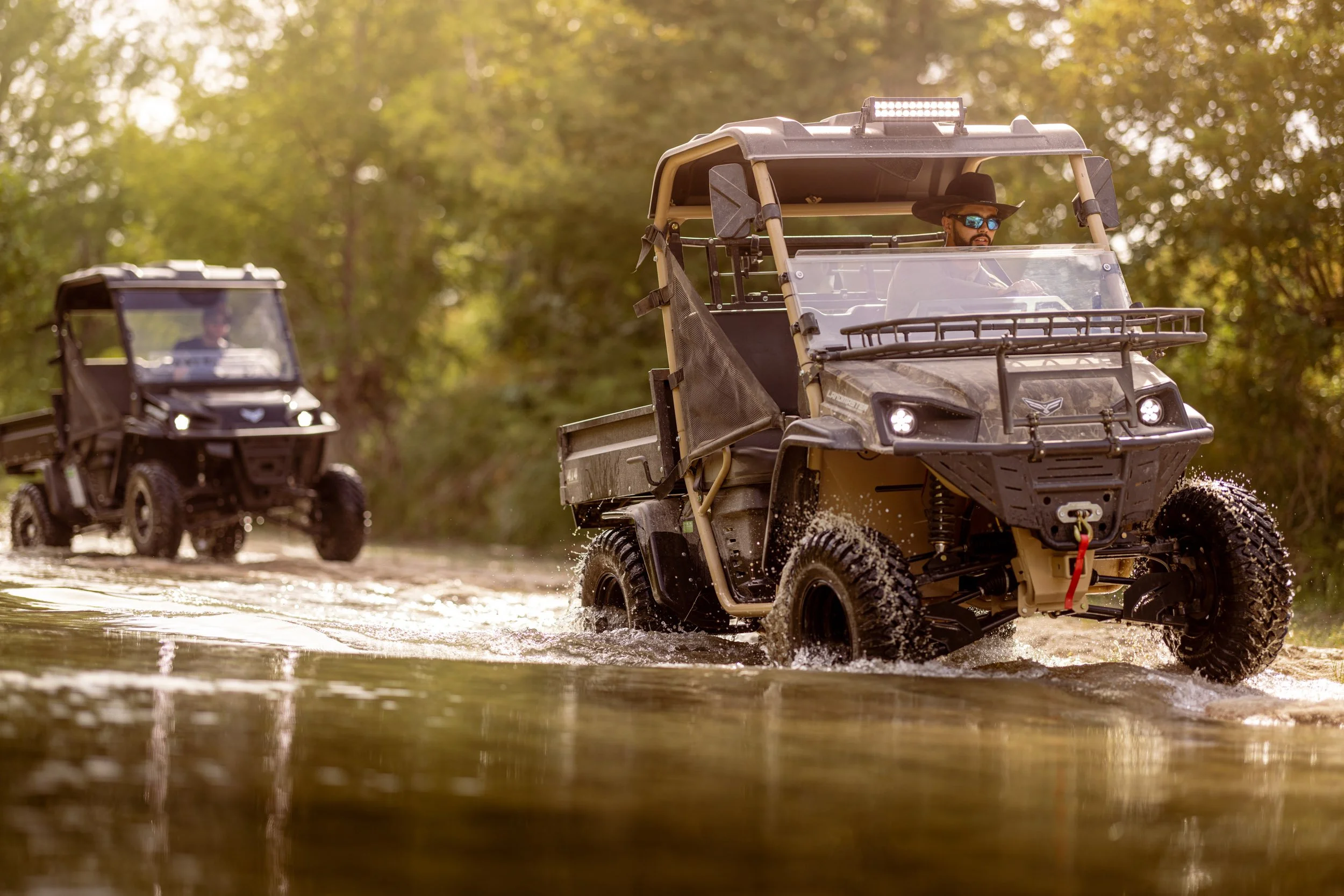 Two all-terrain utility vehicles driving through a shallow water trail in a wooded area during daytime. The front vehicle has a person wearing sunglasses and a black hat, while the second vehicle follows behind.