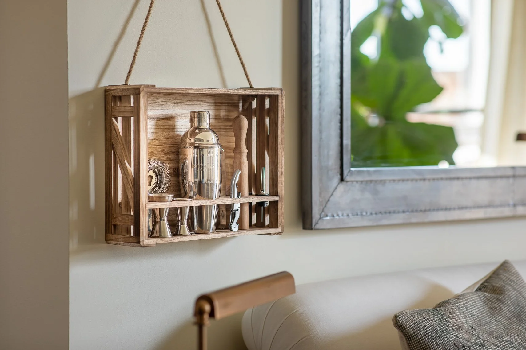 Wooden crate mounted on wall holding metallic cocktail shaker, jigger, and bar tools near a window with greenery outside.