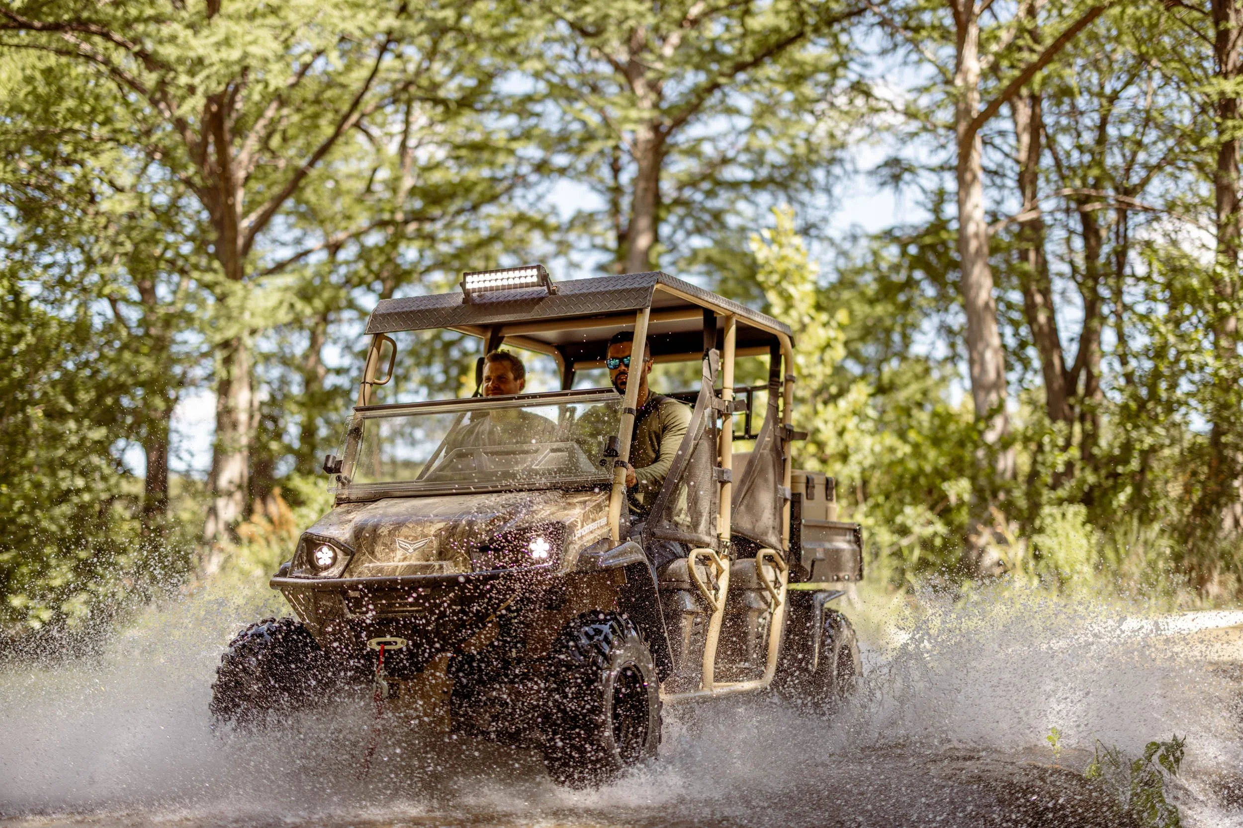 Two men riding an off-road utility vehicle through a shallow water area amidst tall, green trees during daytime.