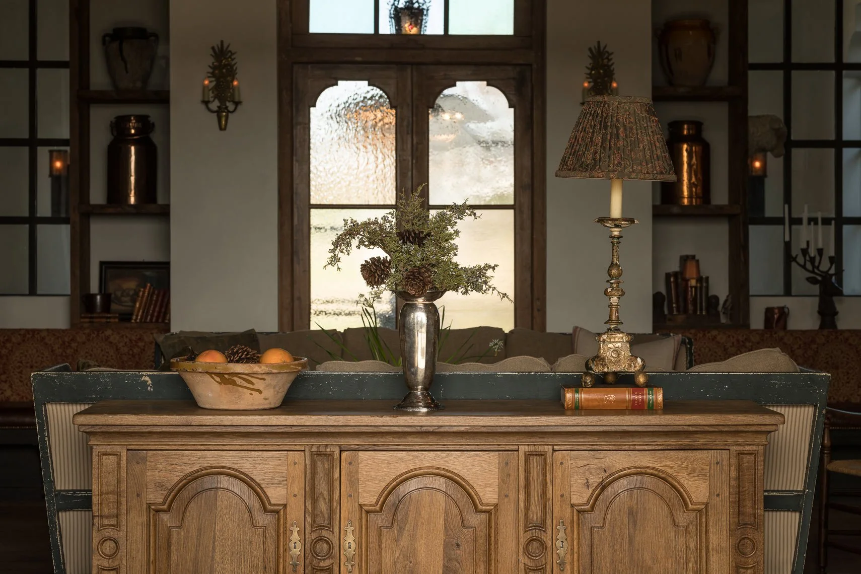 Decorative console table with a floral arrangement in a silver vase, a table lamp with an ornate base and patterned shade, a bowl of apples, and a stack of three books in a cozy living room with wooden shelves and a large window in the background.