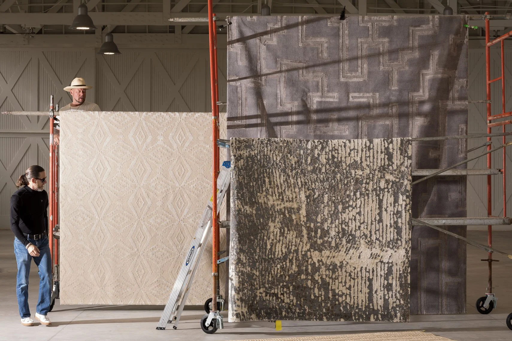 Two men standing near display of carpets hanging on a scaffolding in a showroom or warehouse.