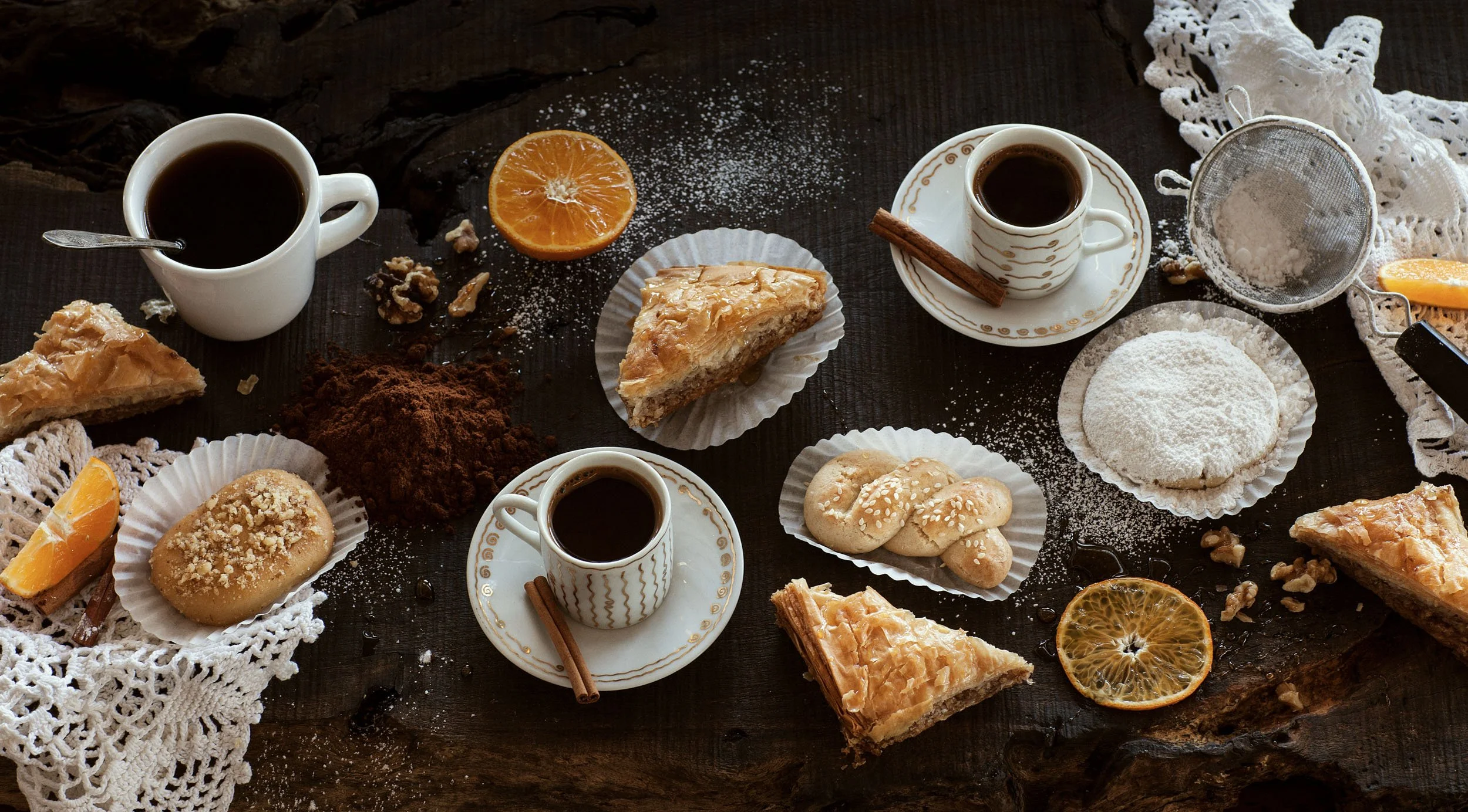 Assorted baked goods and beverages on a rustic wooden table, including coffee cups, slices of pie, cookies, and orange slices, with decorative lace cloths and baking ingredients.