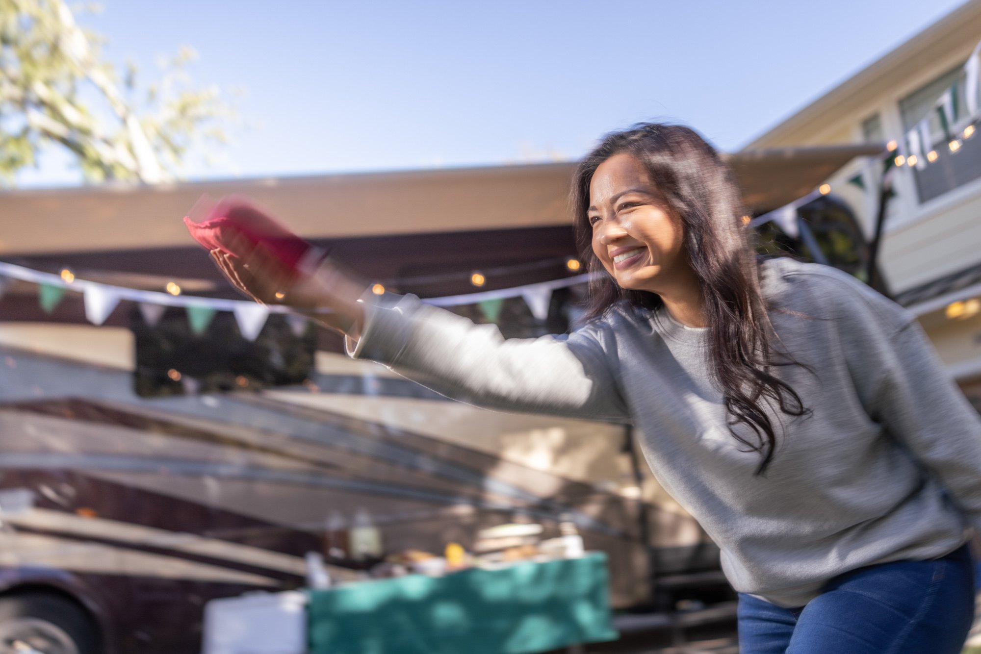 Smiling woman with long dark hair reaching into a container outdoors during daytime, with a house, bunting decorations, and blue sky in the background.