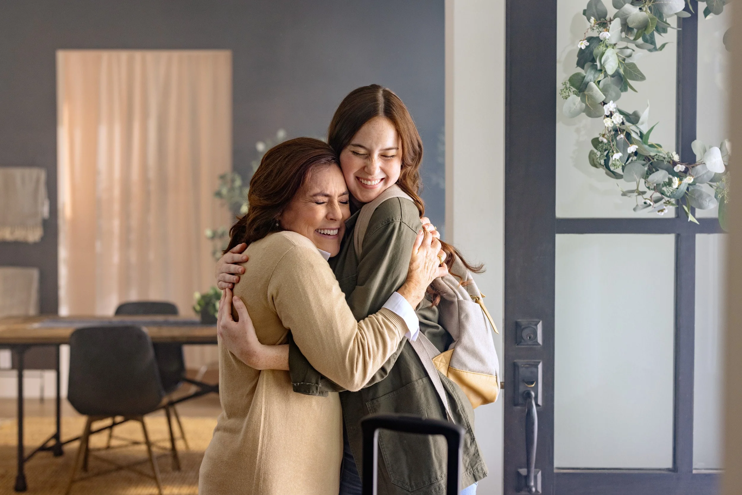 Two women hugging and smiling in a warmly lit room with modern decor and plants.